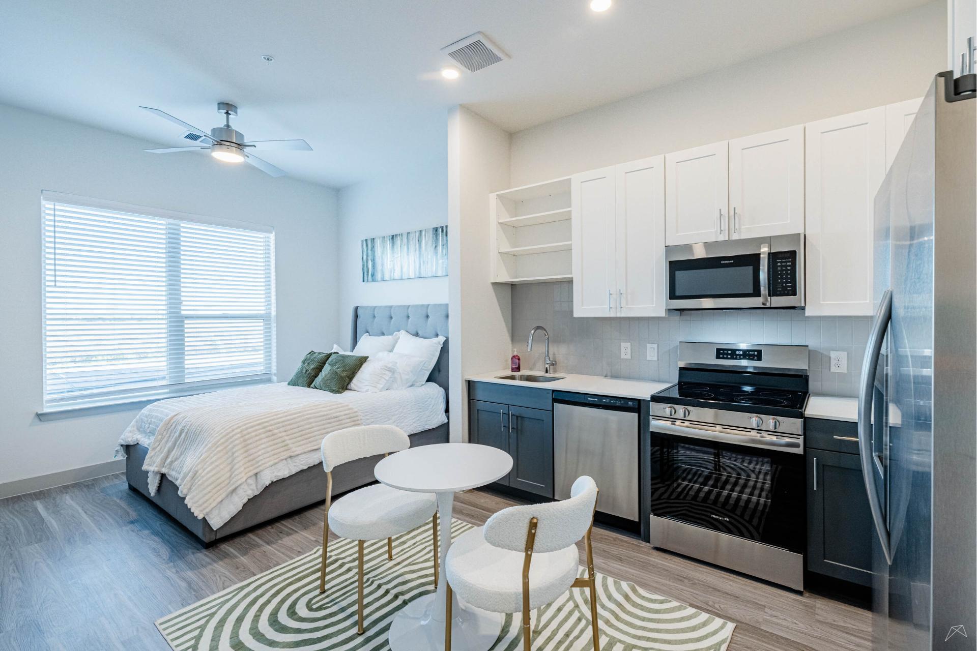 Modern studio apartment with a bed, kitchen area with stainless appliances, white cabinets, small table on rug, large window for natural light.