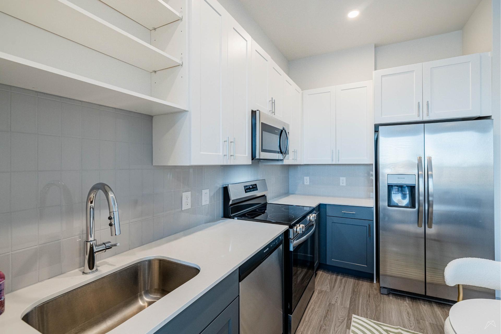 Modern kitchen with white and blue cabinets, stainless appliances, silver sink, light gray tile backsplash, and wood-look flooring.