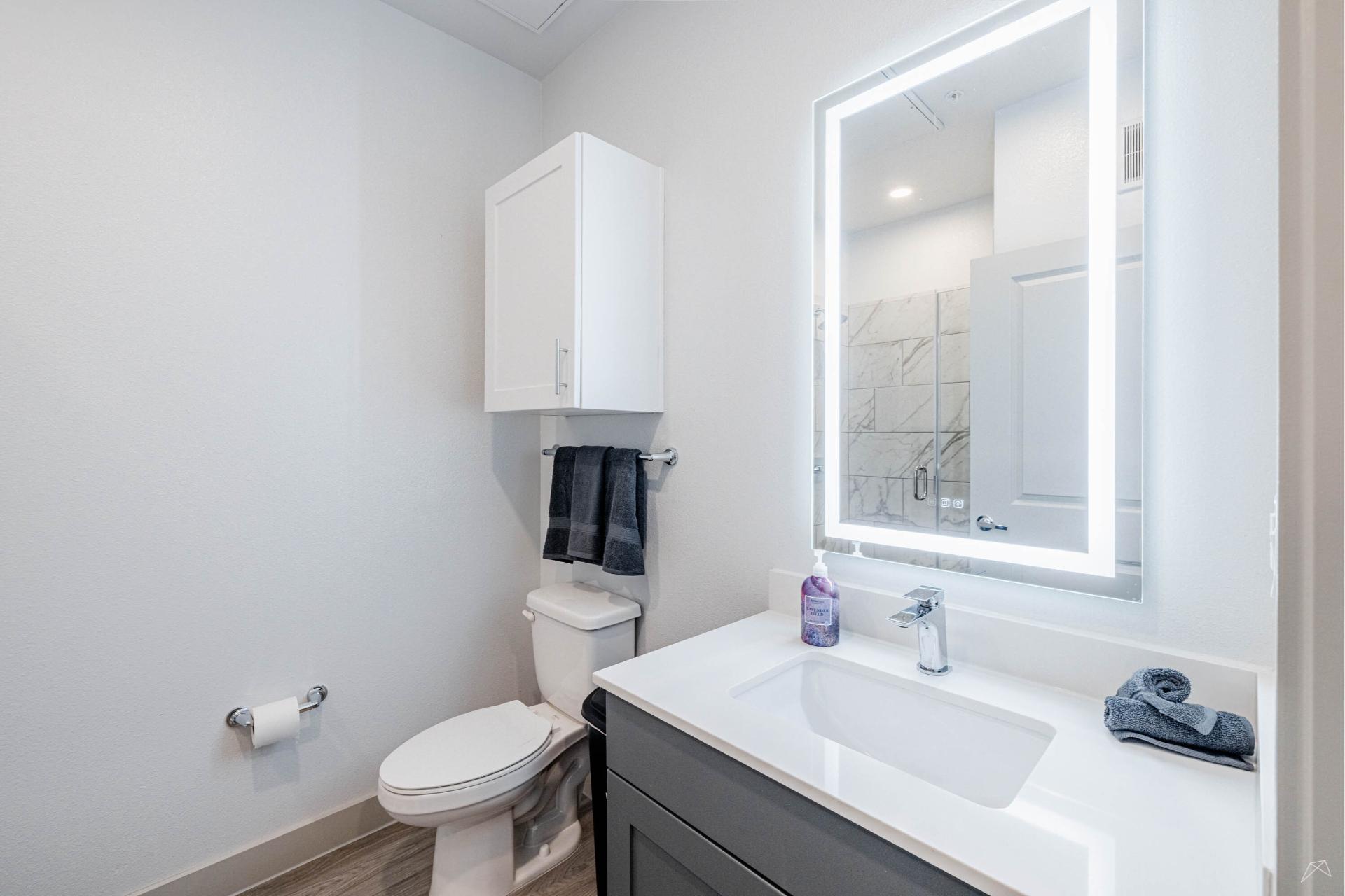 Modern bathroom with white toilet, wall cabinet, wood-look dark vanity with sink, illuminated mirror, towels, and marble-style shower tiles.
