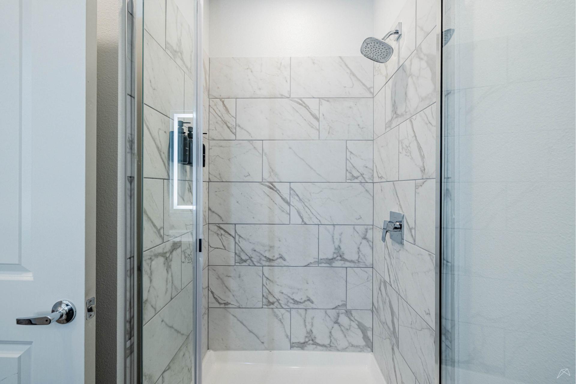 A modern shower with a glass door, marble-patterned tile walls, rainfall showerhead, and a built-in wood-look niche for toiletries.