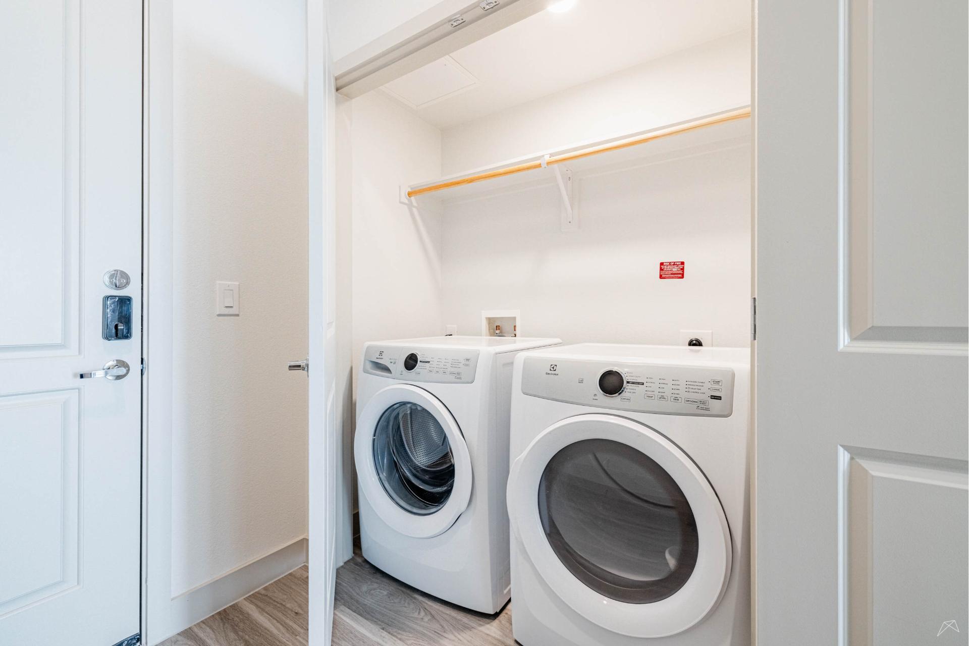 A modern laundry nook with a front-loading washer and dryer side by side, under a wood-look shelf, in a white closet with light flooring.