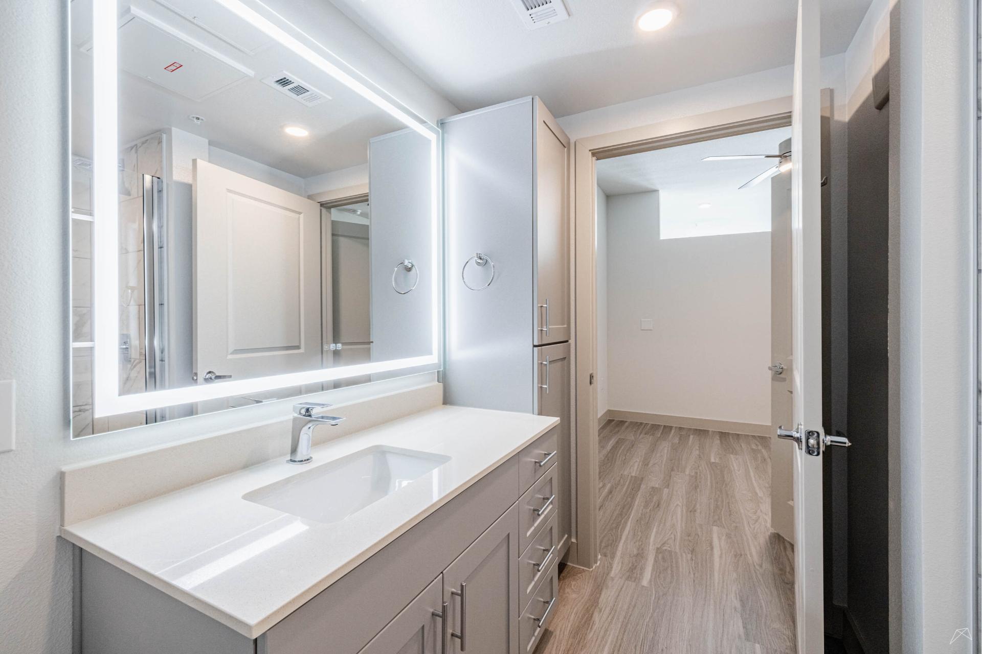 A modern bathroom with a large backlit mirror, white sink, light gray cabinets, wood-look flooring, and an open door to a bright hallway.