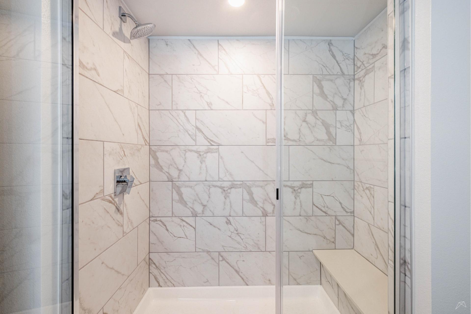 A modern shower with white and gray marble tile walls, a glass door, rainfall showerhead, wood-look bench on the right, chrome faucet.