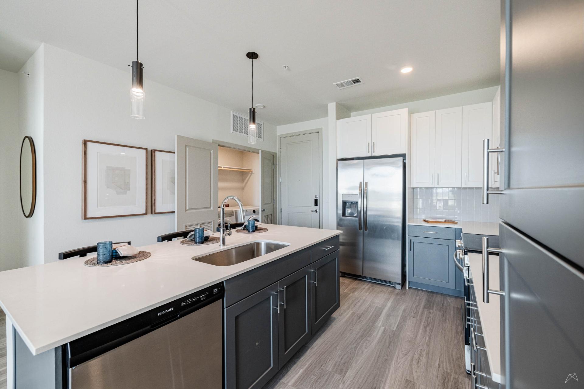 Modern kitchen with a large island, sink, stainless appliances, two-tone cabinets, pendant lights, and light wood-look flooring.