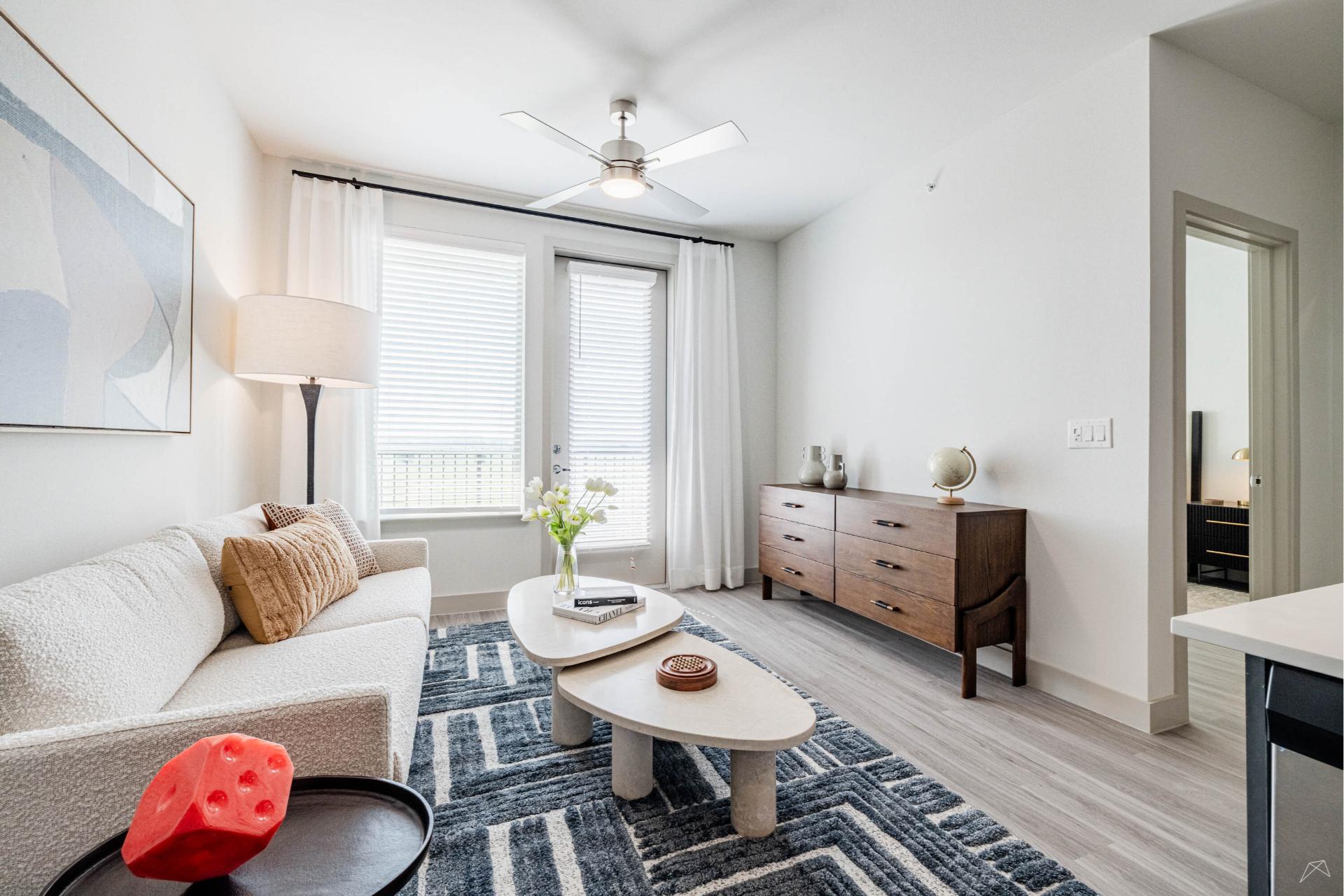 A modern living room with a white sofa, abstract art, two oval coffee tables, geometric rug, wood-style dresser, and sheer curtains.