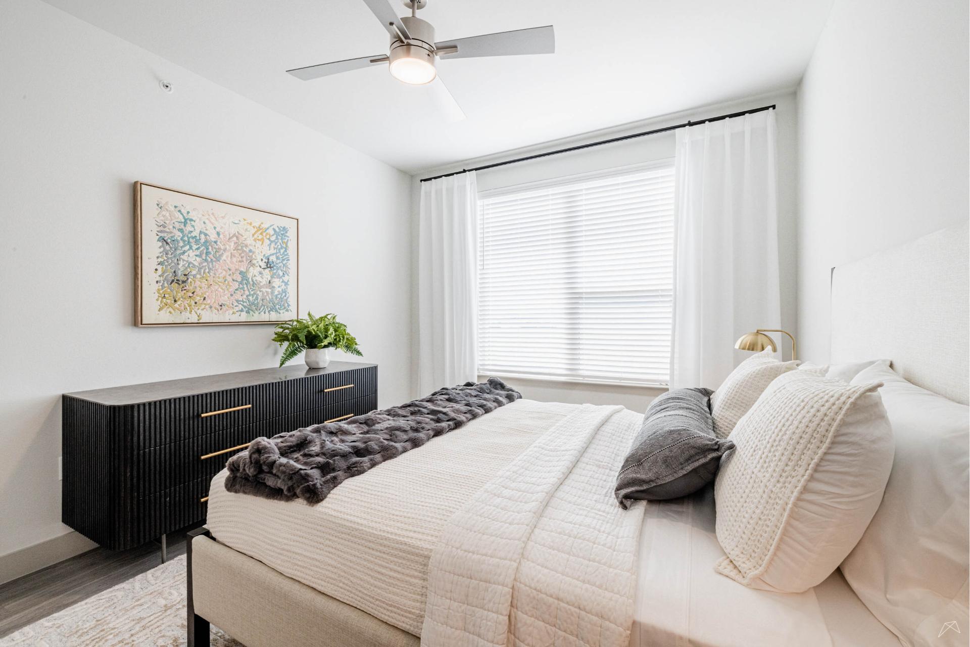A bright, modern bedroom with a large window, white walls, neutral bedding, a dark wood-look dresser with a plant and art above.