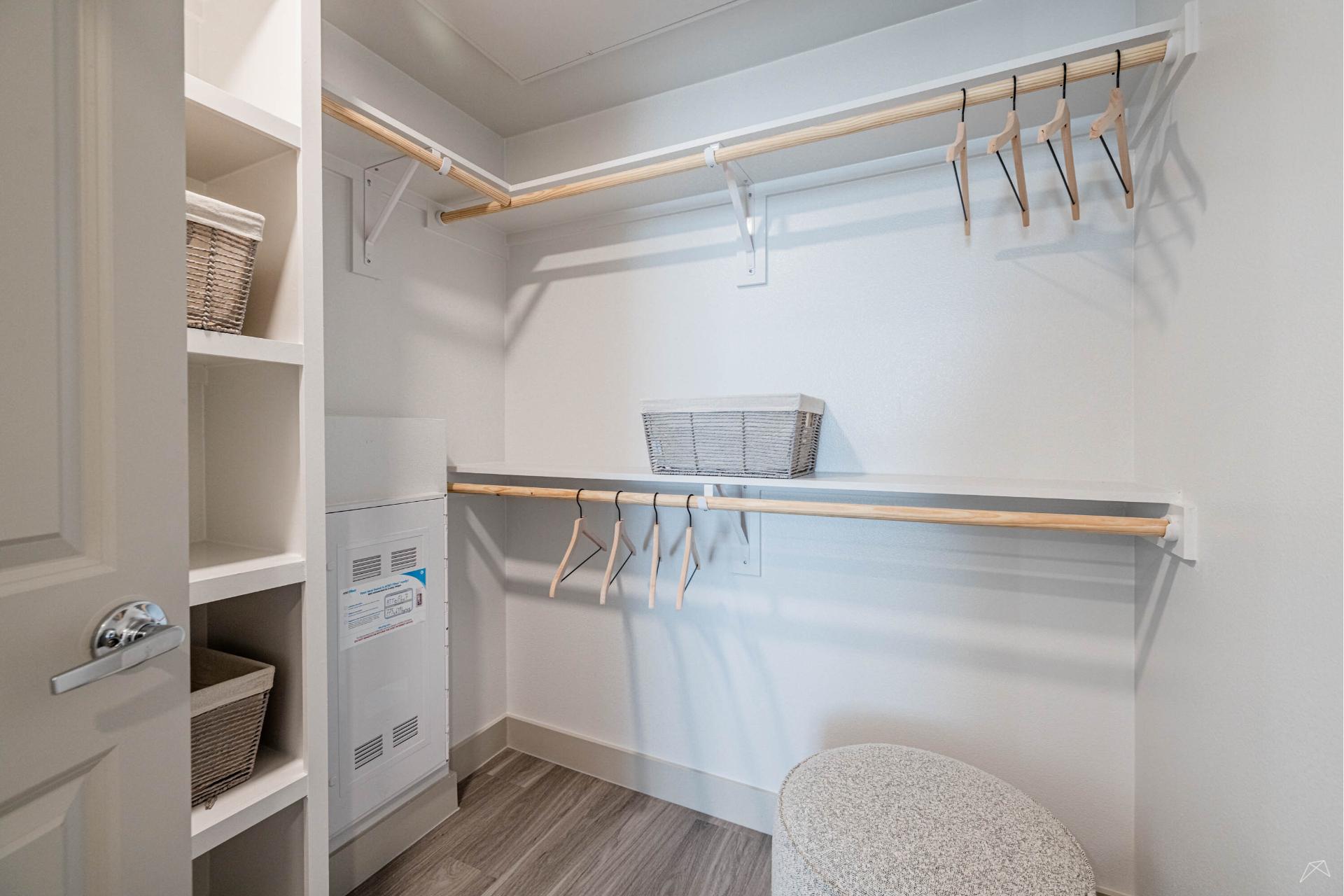 A spacious walk-in closet with wood-style rods, empty hangers, open shelves and baskets, a lower basket, and a round padded stool on a wood-style floor.