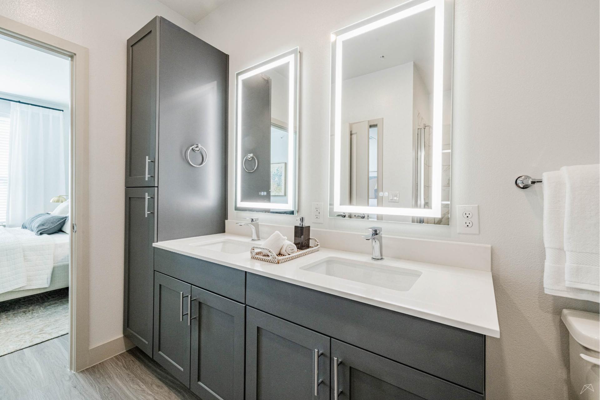 Modern bathroom with dual sinks, gray cabinets, illuminated mirrors, white countertops, wood-look tall storage cabinet, towel on right.