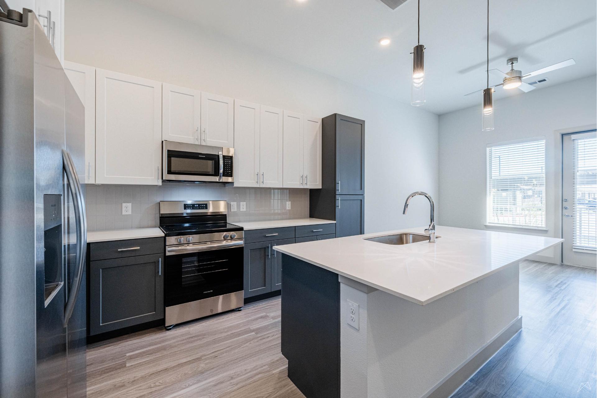 Modern kitchen with white and dark gray cabinets, stainless steel appliances, granite-style island with sink, pendant lights, and large windows.