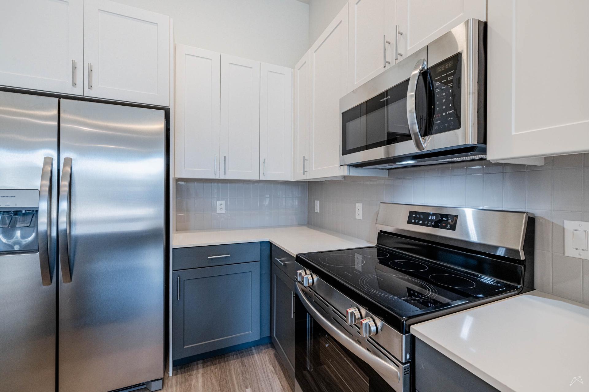 Modern kitchen with stainless steel appliances, white uppers, dark lowers, light countertops, gray tile backsplash, and wood-look flooring.