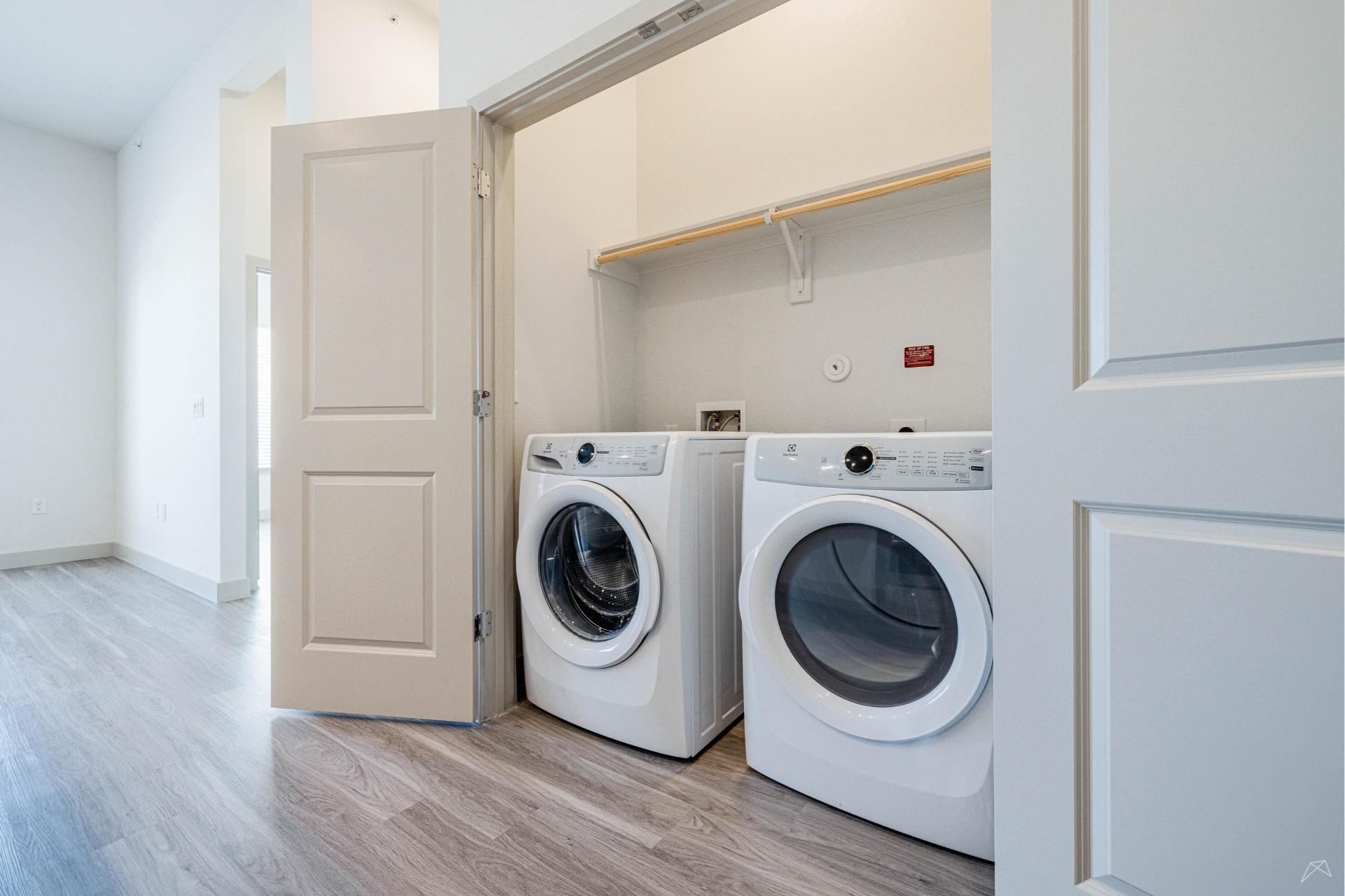 A laundry area with front-loading washer and dryer, white folding doors open, a shelf above, in a room with wood-look flooring and white walls.