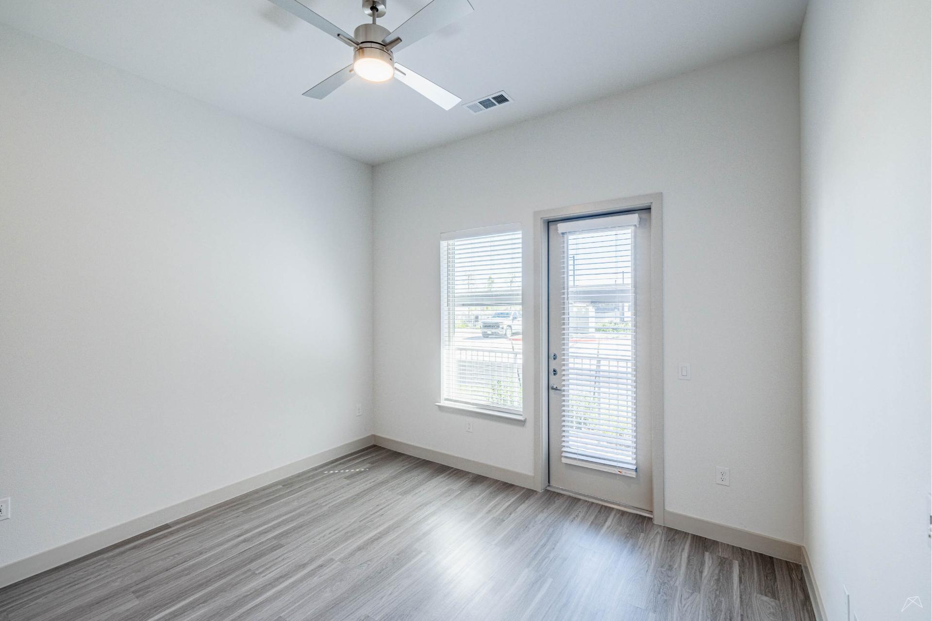 A bright, empty room with light gray wood-look flooring, white walls, ceiling fan with light, window, glass door with blinds. Natural light fills the space.