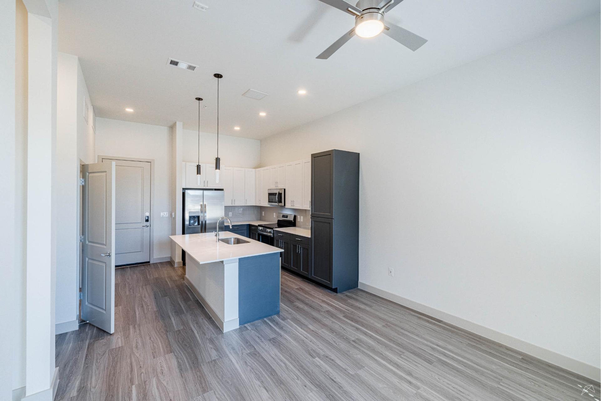 Modern kitchen with gray cabinets, stainless steel appliances, large island with sink, pendant lights, wood-look flooring, white walls.