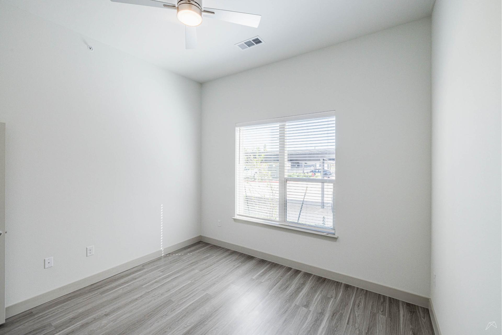 A bright, empty room with light gray wood-look flooring, white walls, a big window with blinds, and a ceiling fan lit by sunlight.