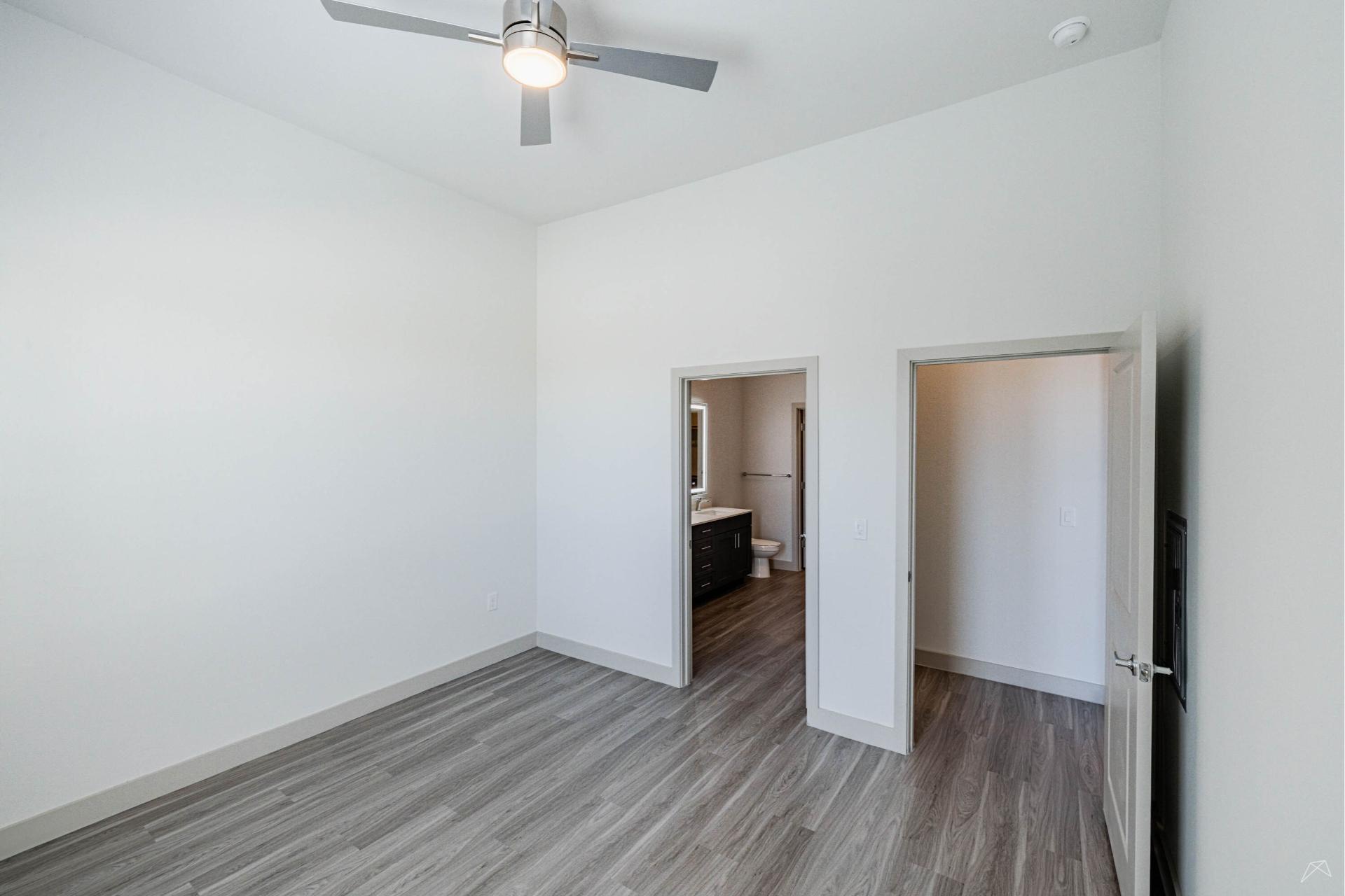 Minimalist, empty bedroom with light gray wood-look flooring, white walls, a ceiling fan, and two doors—one to a bath, one to another room.