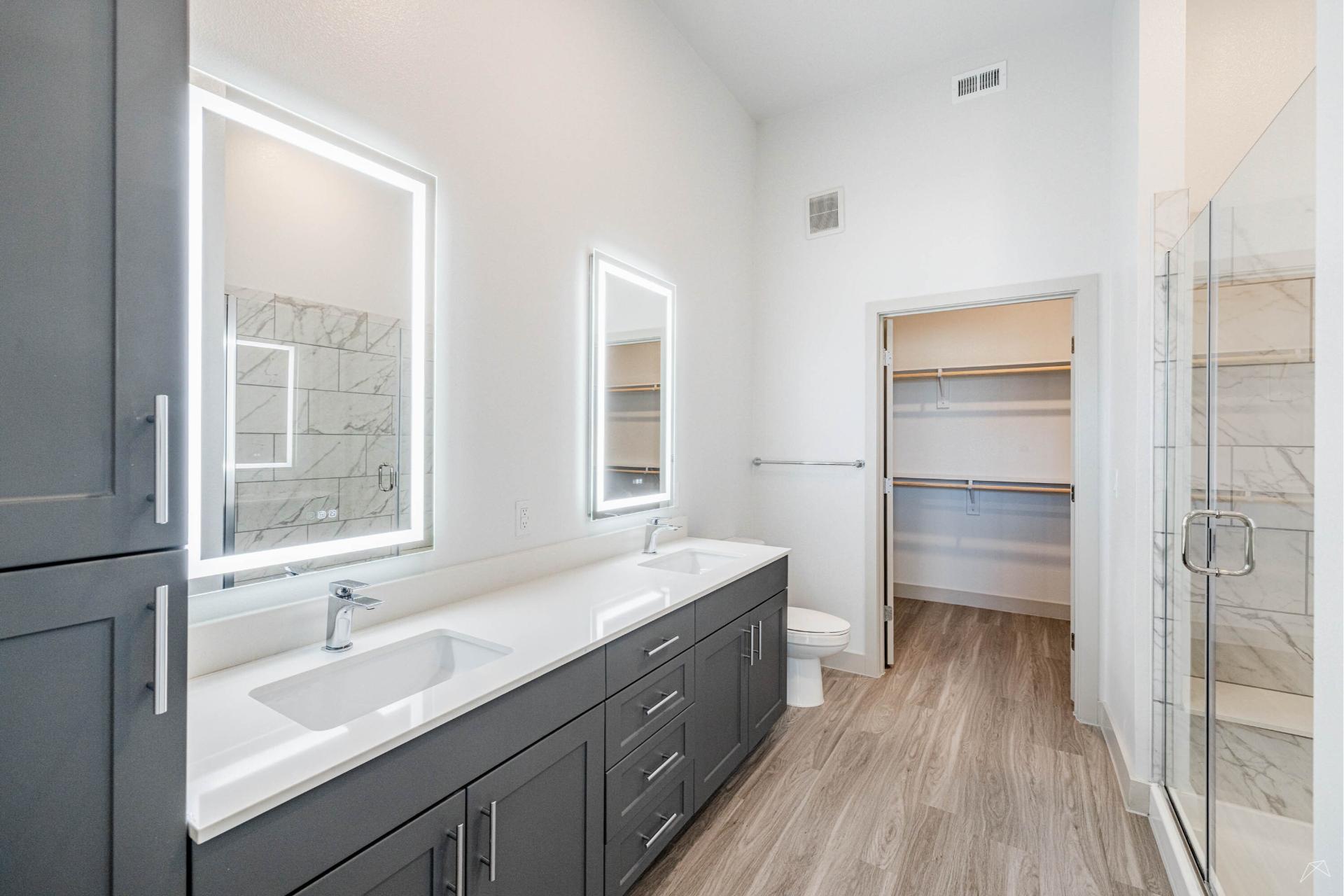 Modern bathroom with double sinks, large mirror cabinets, glass shower, light wood-look flooring, toilet, and spacious walk-in closet.