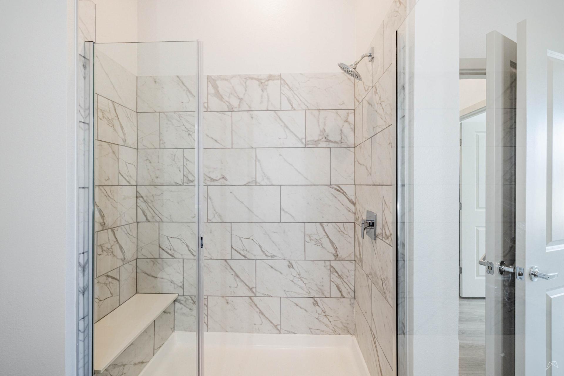 A modern shower with large white marble tiles, glass door, built-in bench on the left, chrome fixtures; open doorway outside.