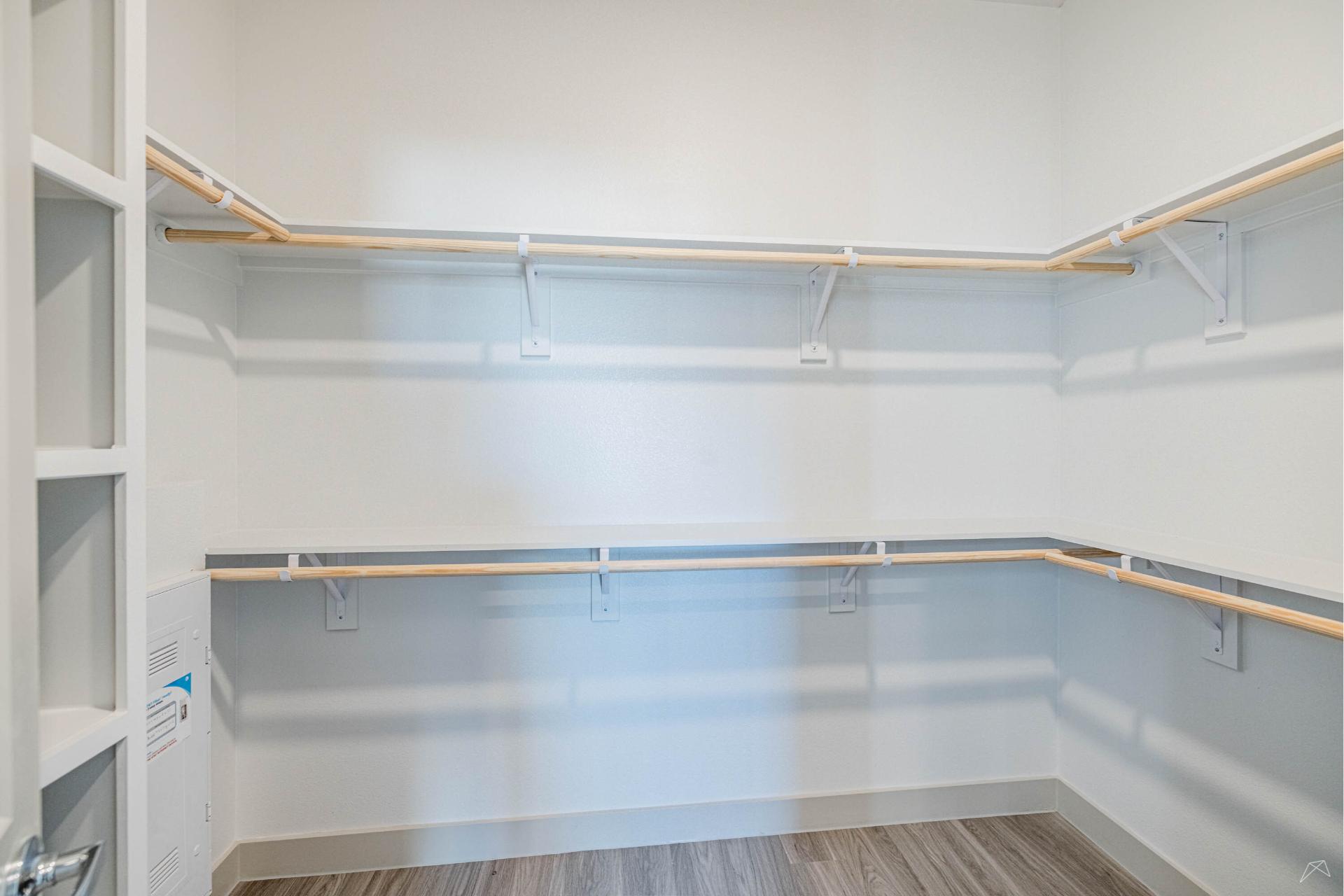 Empty walk-in closet with white walls, light wood-look flooring, and white shelves with wood-like rods on three sides for hanging clothes.