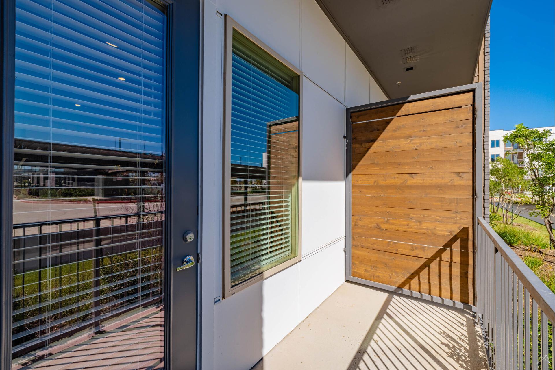 A modern apartment balcony with a wood-look privacy screen, metal railing, and glass door and window with closed blinds, overlooking a sunny outdoor area.