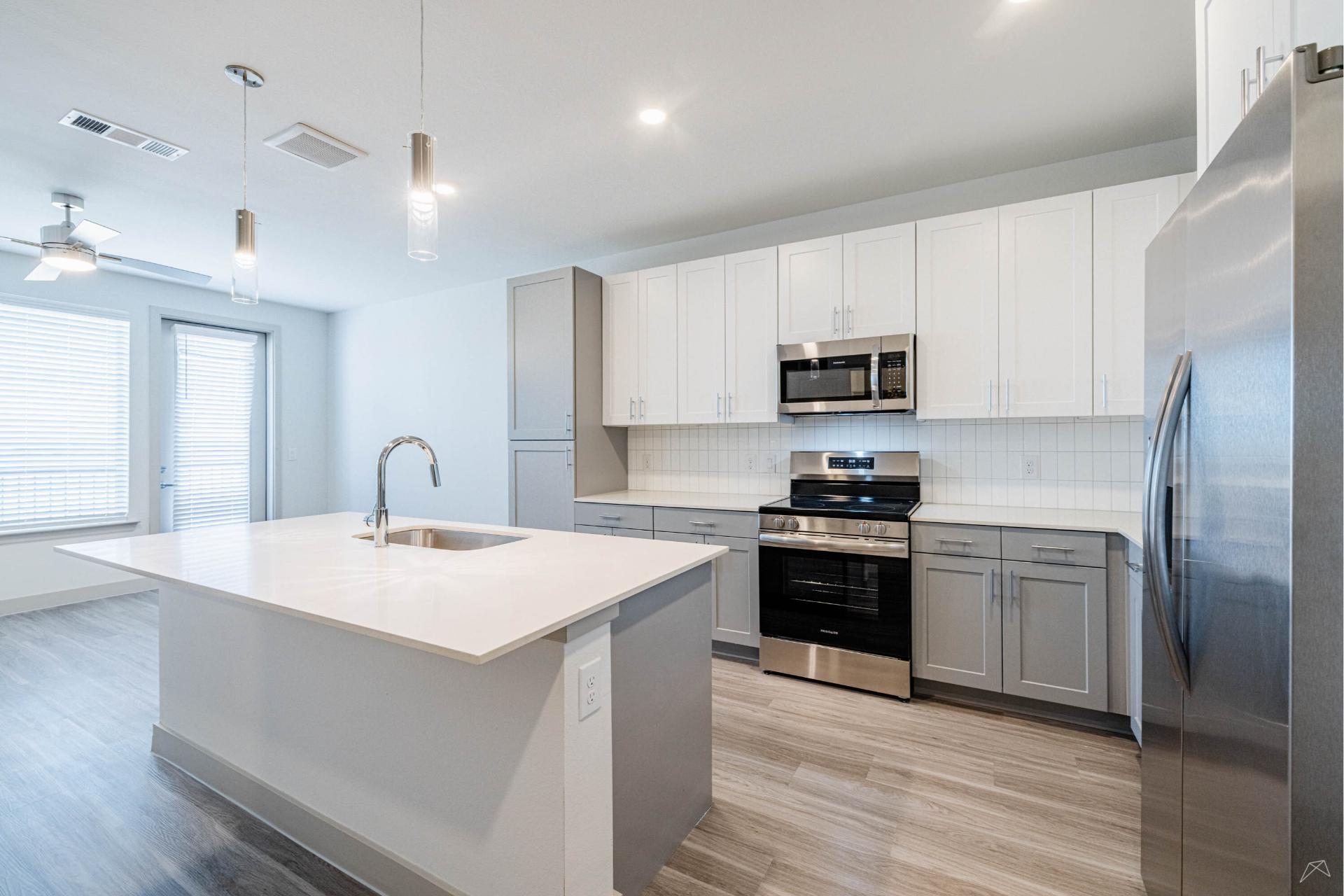 Modern kitchen with stainless steel appliances, white and gray cabinets, large island with sink, pendant lights, wood-style flooring, bright from big windows.