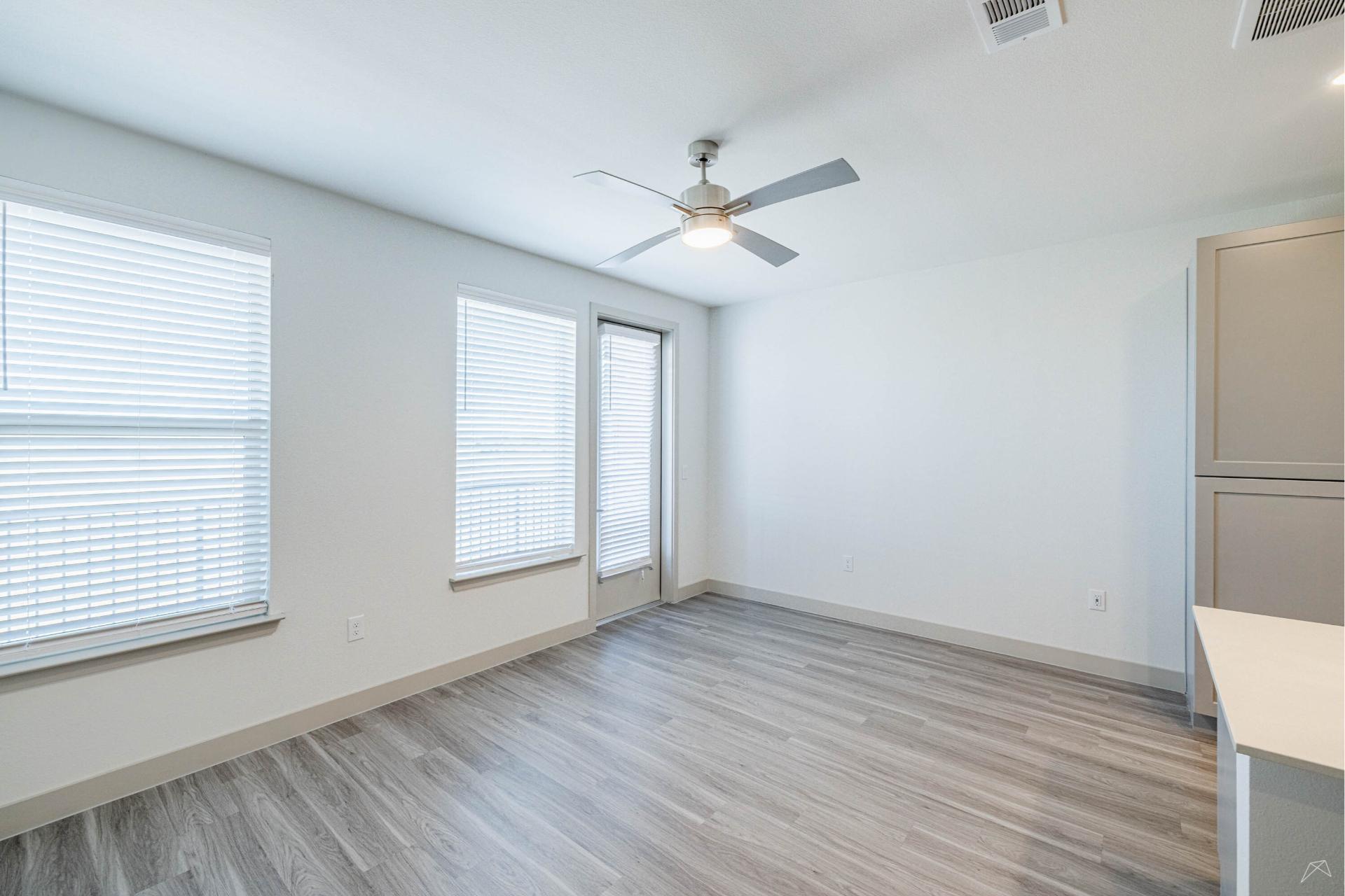 Bright, empty room with light gray wood-look flooring, white walls, big windows with blinds, ceiling fan, glass door out, and kitchen counter.