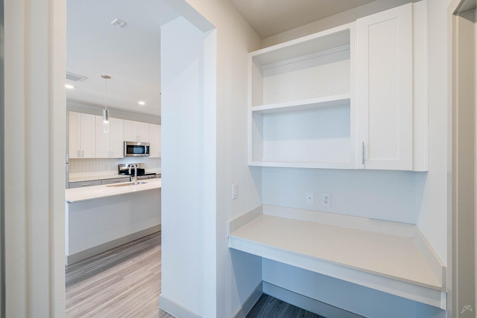 Small built-in desk nook with white cabinets and open shelves, quartz countertop, outlets, off a hall leading to a modern kitchen.