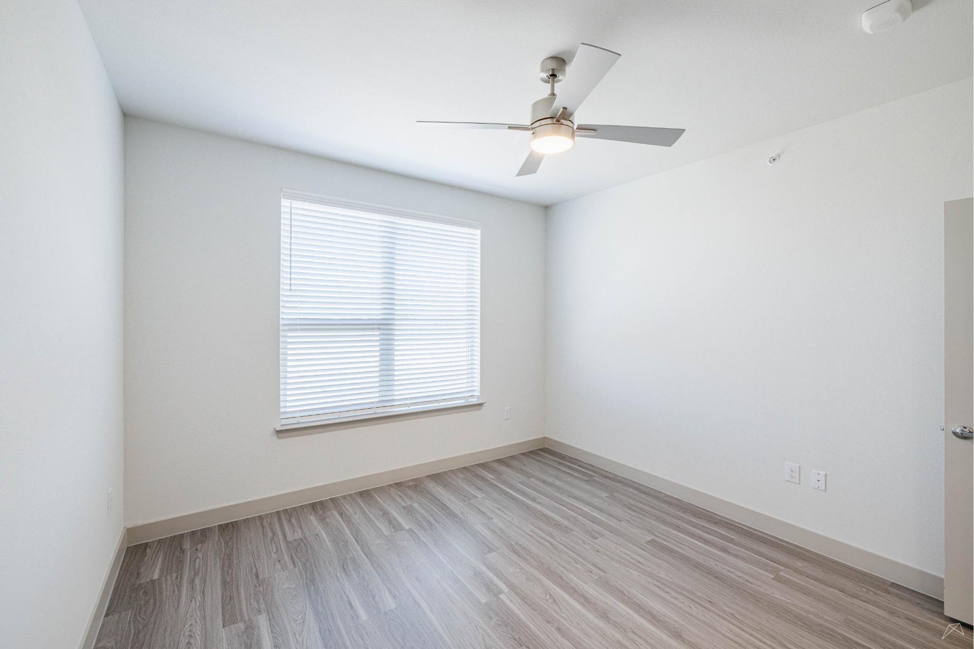 A bright, empty room with light-colored walls, large window with blinds, wood-look flooring, and a ceiling fan with a light fixture.