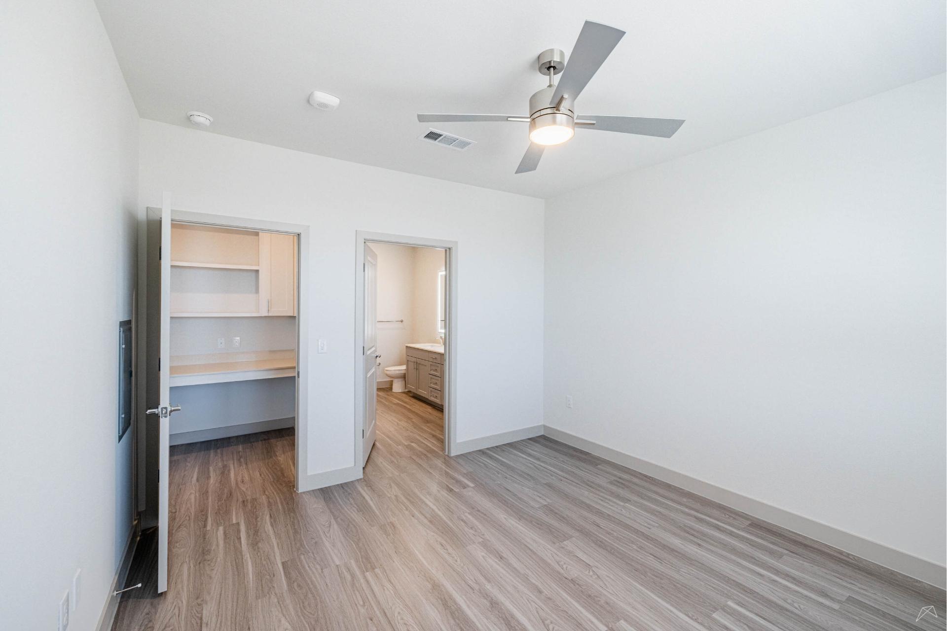 A bright, unfurnished room with light gray wood-like flooring, white walls, a ceiling fan, a closet, and an open door to a bathroom.