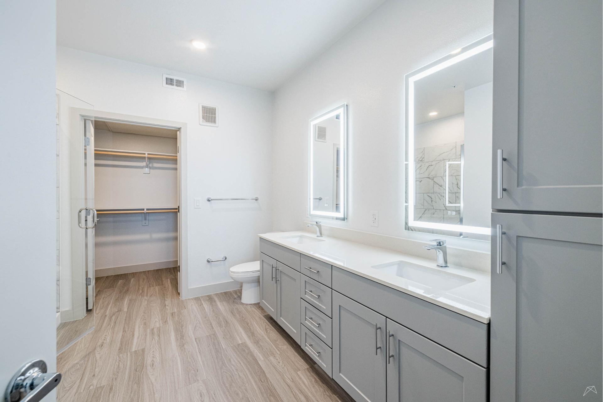 Modern bathroom with light wood-look flooring, gray double-sink vanity, backlit mirrors, open closet with shelves, and white walls.