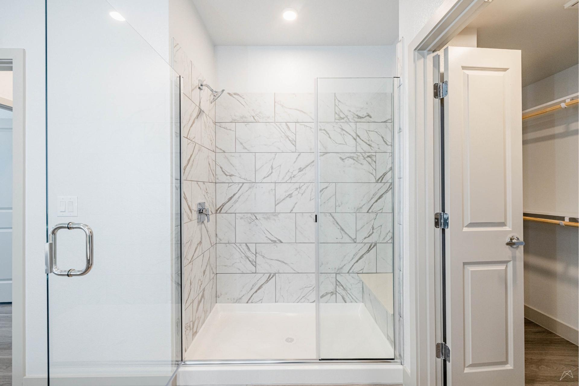 Modern bathroom with a large glass shower, white and gray marble tiles, next to an open walk-in closet with wood-look shelves and rods.