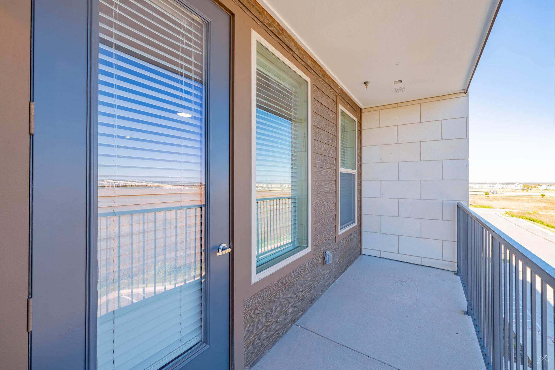 A modern balcony with a glass door, two large windows, beige siding, metal railing, and concrete floor, featuring wood-look accents.