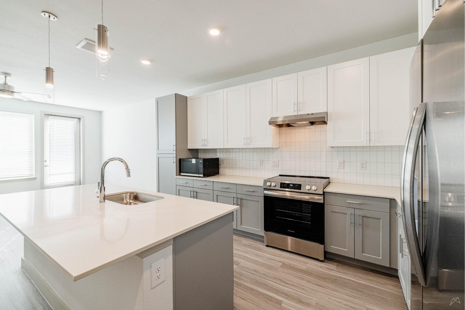 Modern kitchen with white and gray cabinets, stainless steel appliances, island with sink, built-in microwave, and light wood-look flooring. Bright and spacious with natural light from windowed doors.
