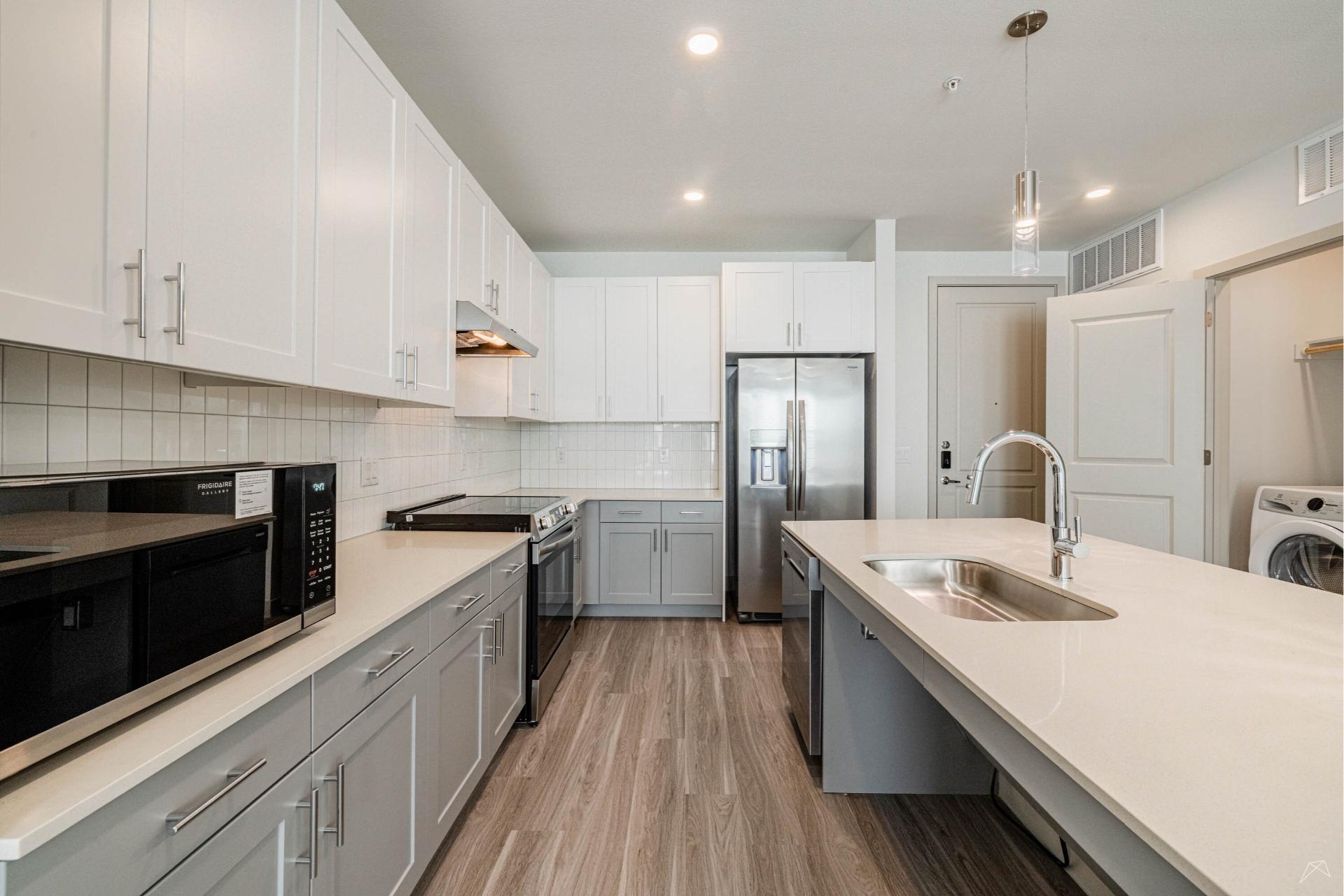 Modern kitchen with white cabinets, stainless steel appliances, large island double sink, laundry area, and wood-look flooring throughout.