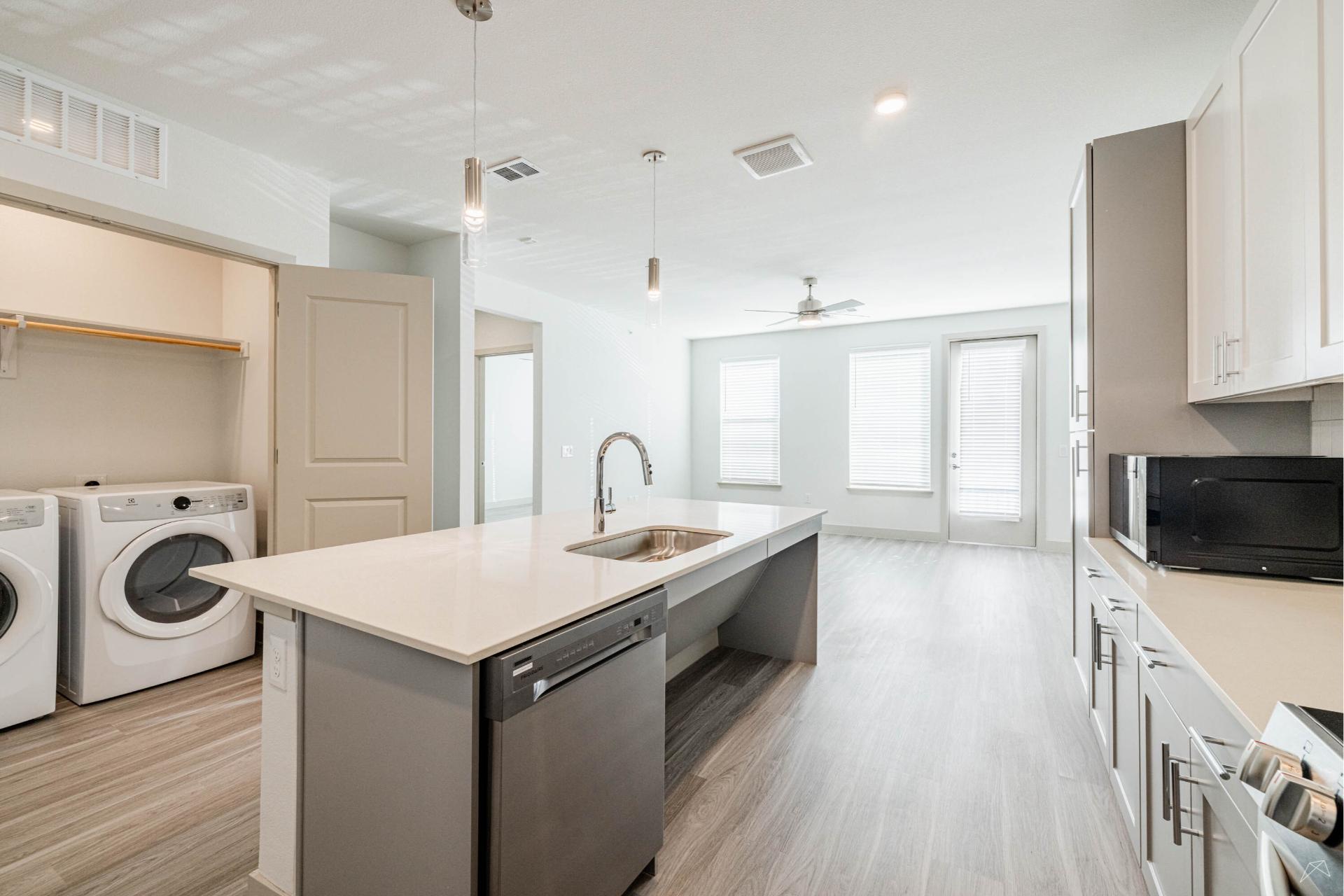 Bright, modern kitchen with a large island, stainless steel appliances, white cabinets, and laundry area. Open to living space with wood-like floors.
