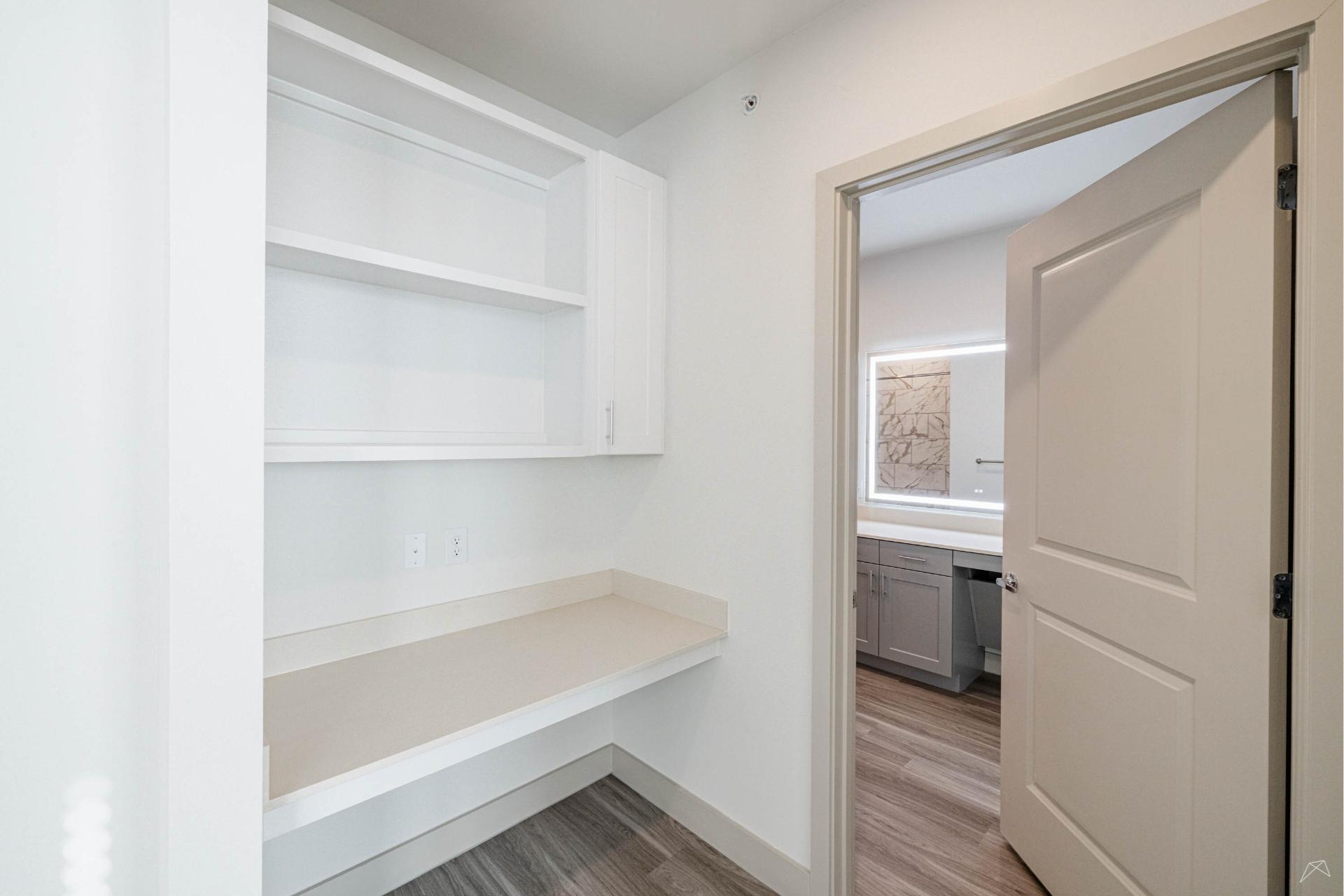 A small, clean room with built-in white shelves, a beige countertop, and wood-like floors leads to a bathroom with vanity and large mirror.
