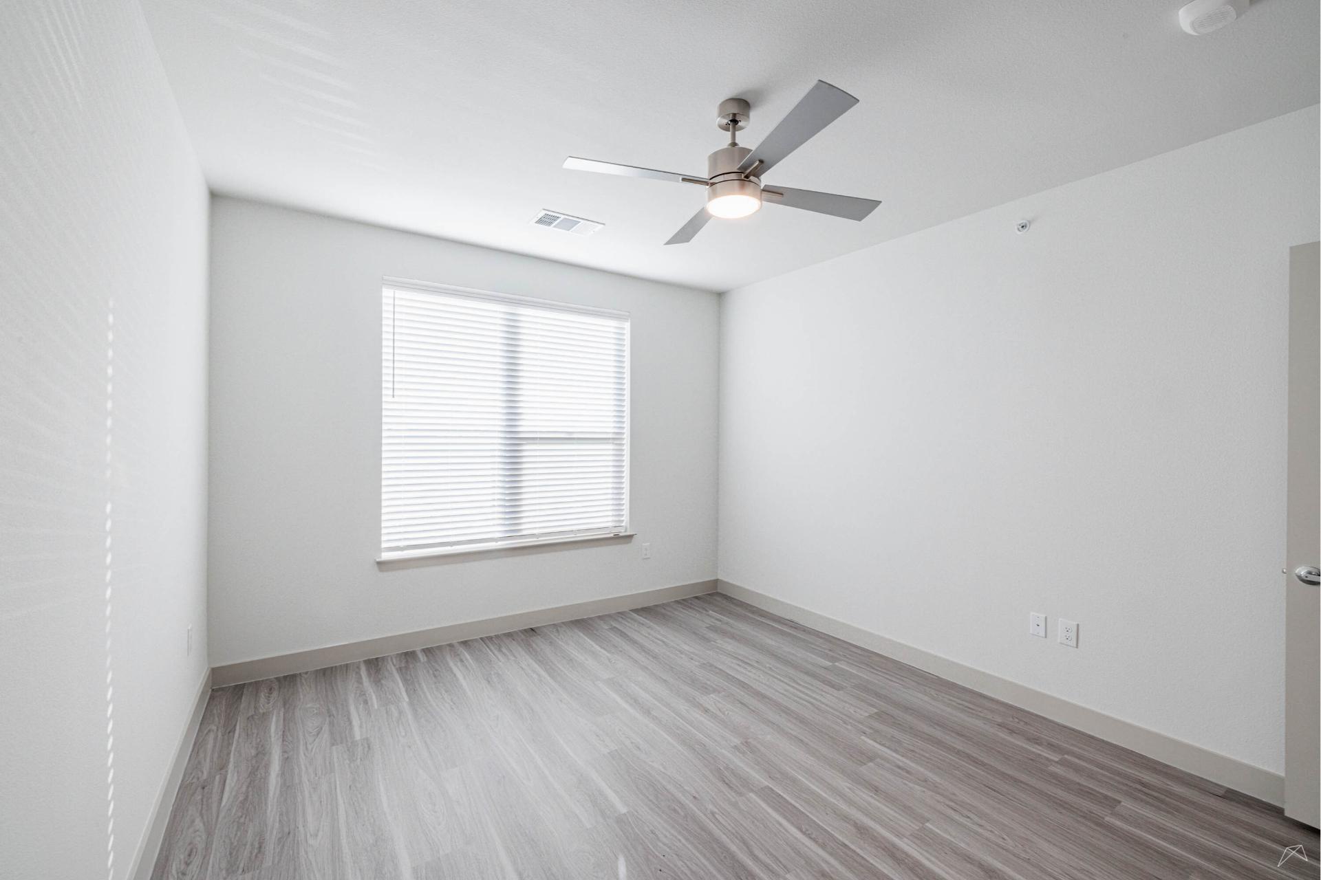 A bright, empty room with light gray wood-look flooring, white walls, a large window with closed blinds, ceiling fan, and open door.