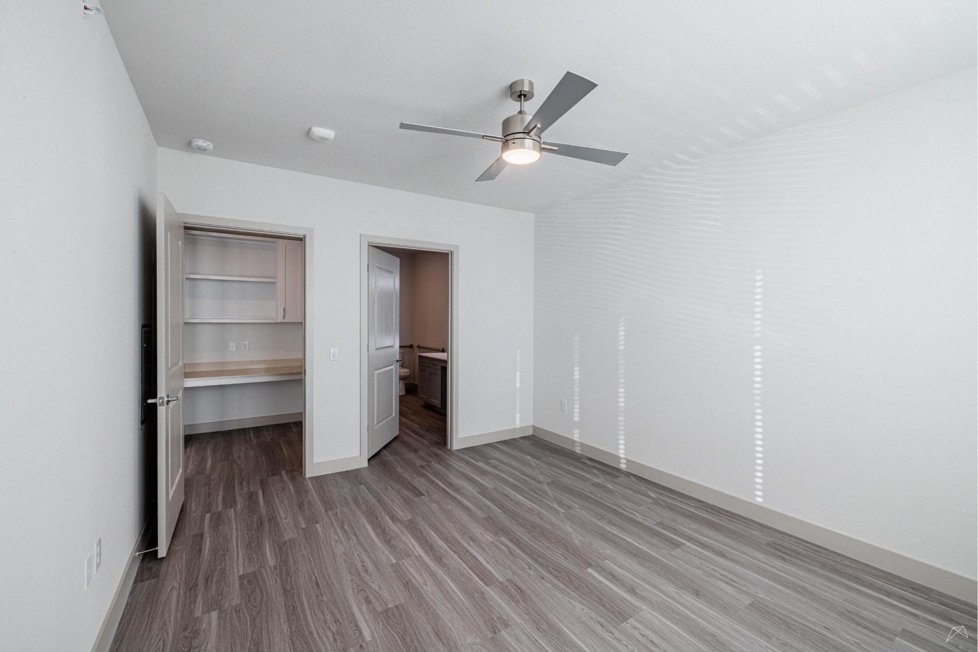Empty modern room with gray wood-look floors, white walls, ceiling fan with light, open doorways to a desk area and bathroom, sunlight streaming in.