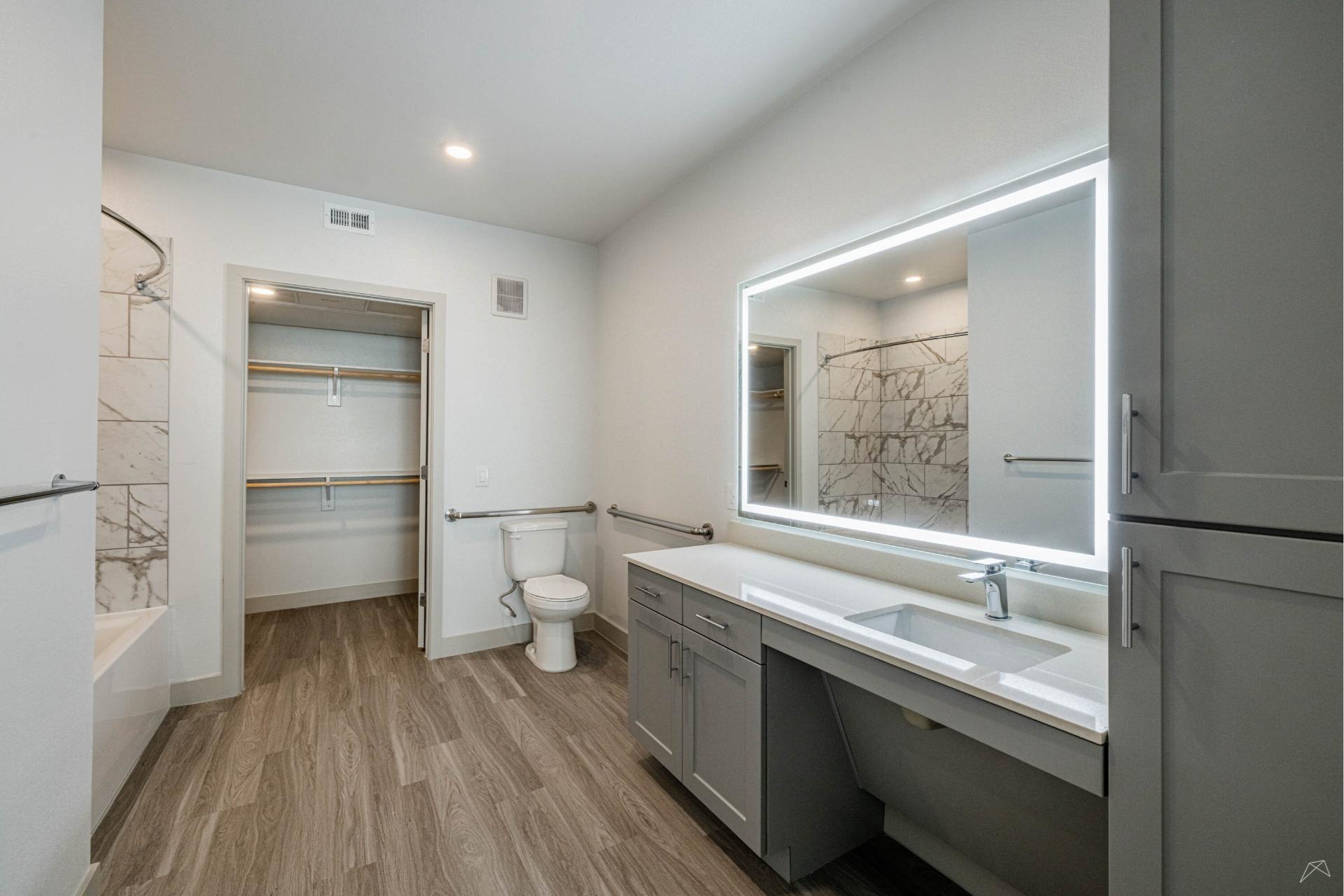 Modern bathroom with wood-look flooring, large lighted mirror above gray vanity, toilet, open closet, and marble-patterned shower wall.