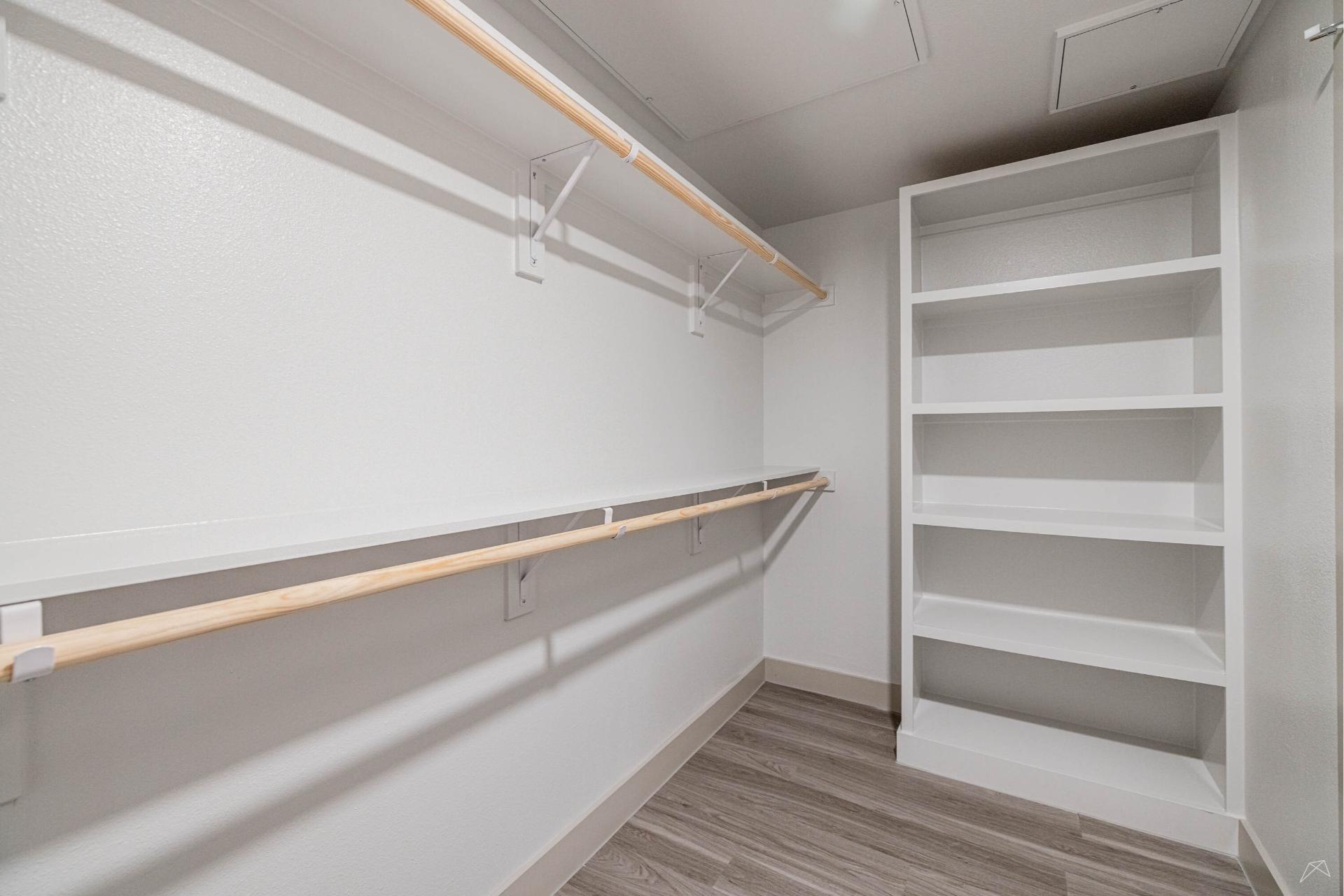 A spacious empty walk-in closet with white walls, rods for hanging clothes, open shelving units, and wood-like flooring.