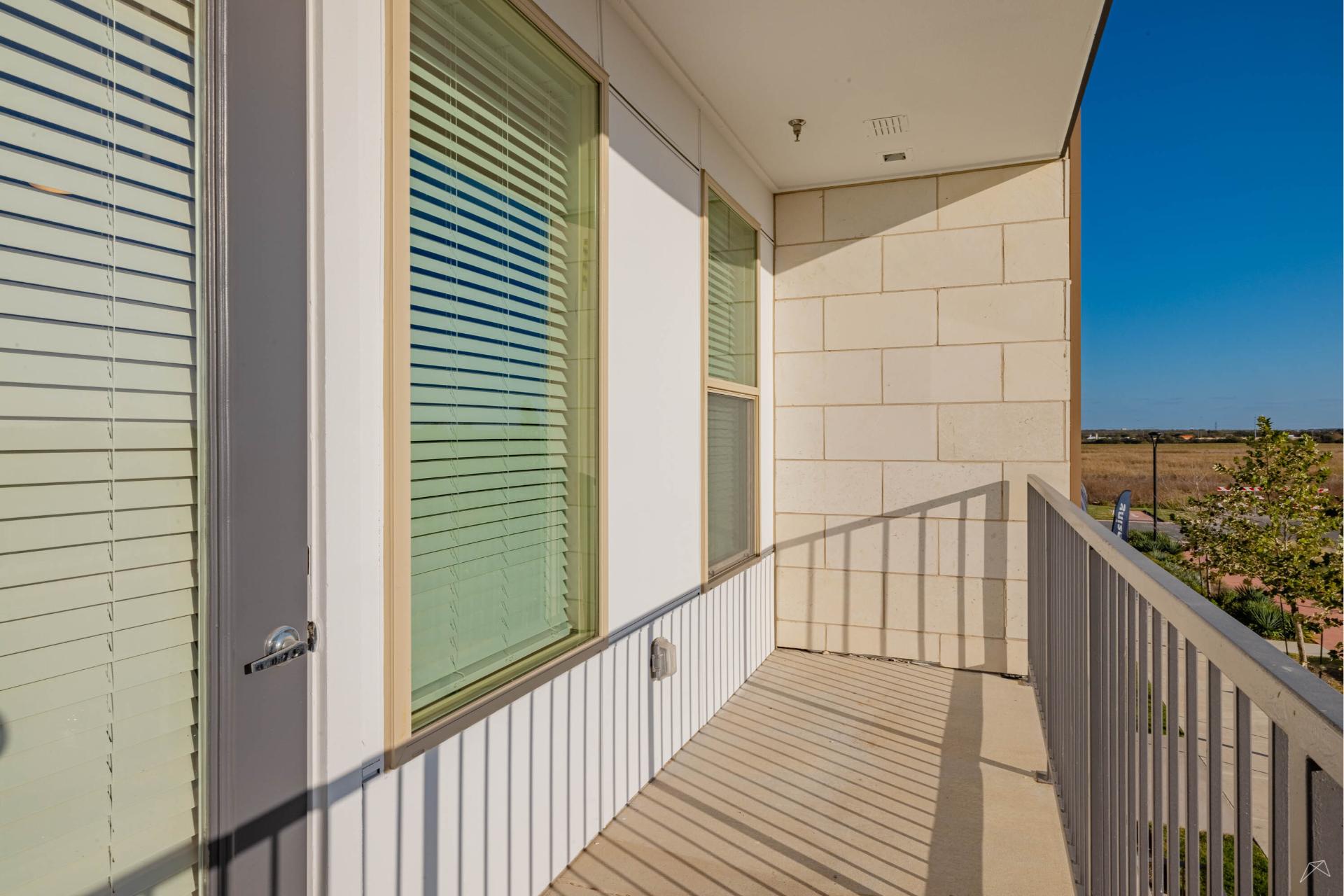 A sunny balcony with beige stone walls, metal railing, and two windows with closed blinds, overlooking a field and blue sky.