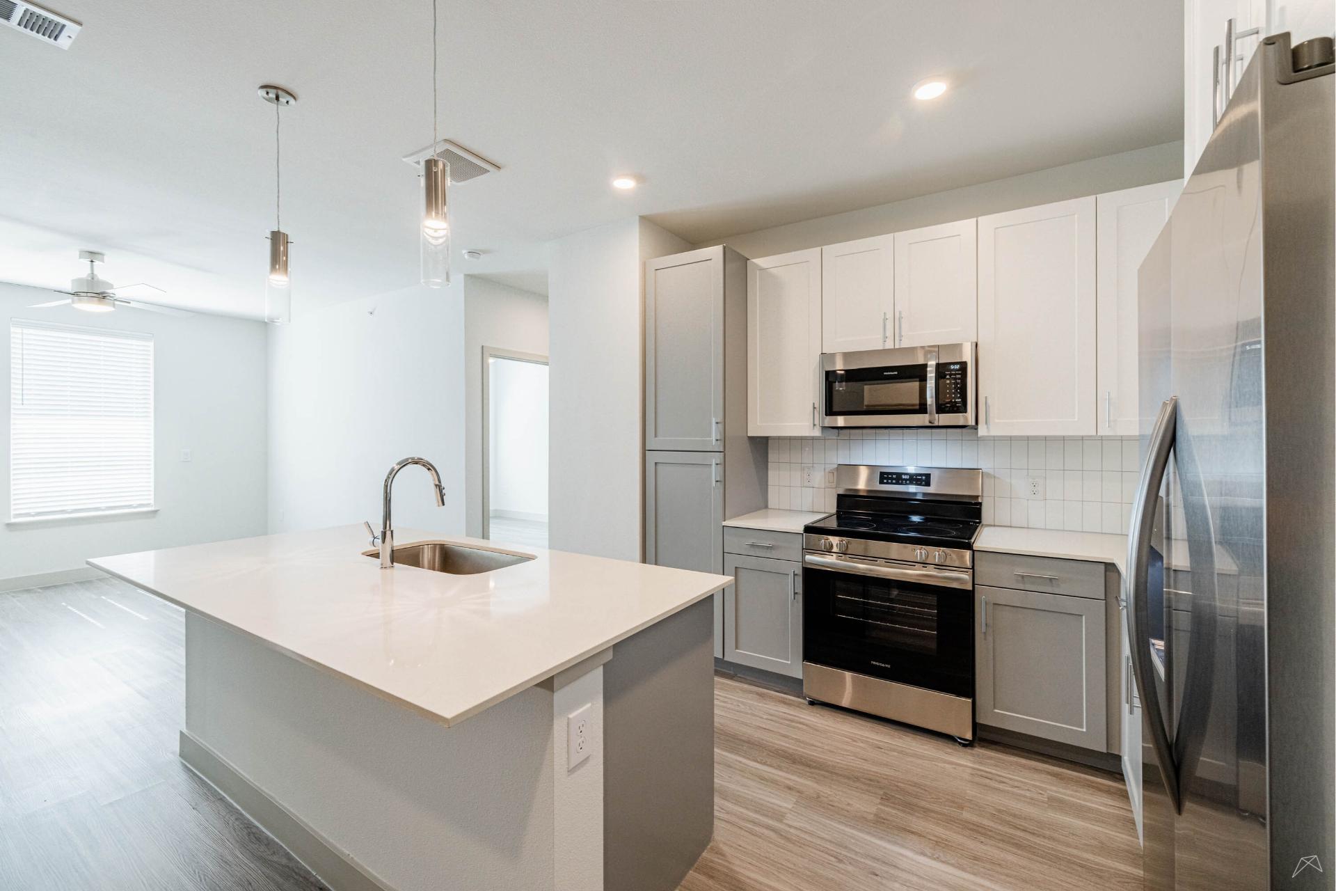 Modern kitchen with white cabinets, stainless appliances, an island with sink and pendant lights, and light wood-look flooring. Open living area nearby.