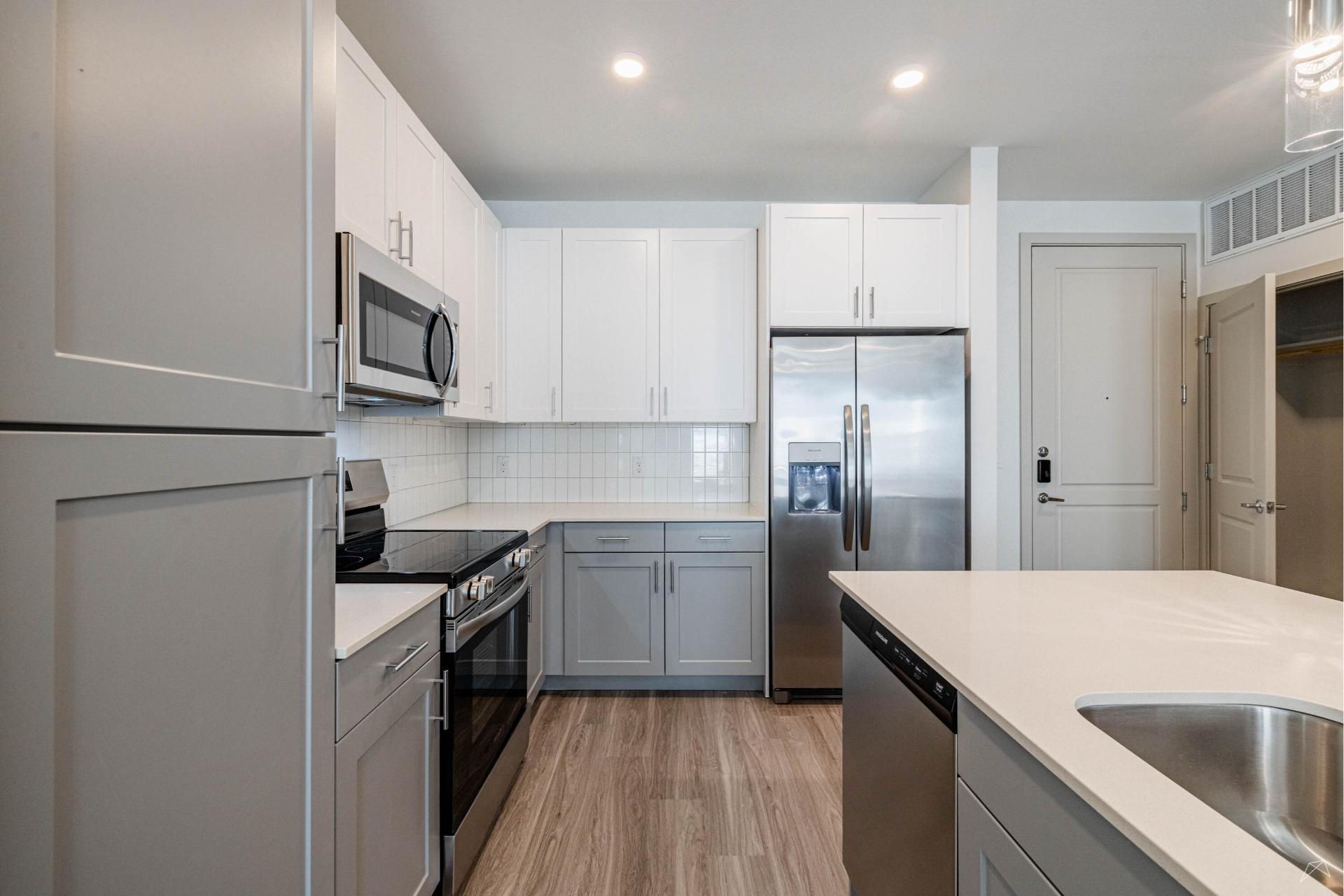 Modern kitchen with light gray cabinets, stainless steel appliances, white tile backsplash, and wood-style flooring. Island with sink.