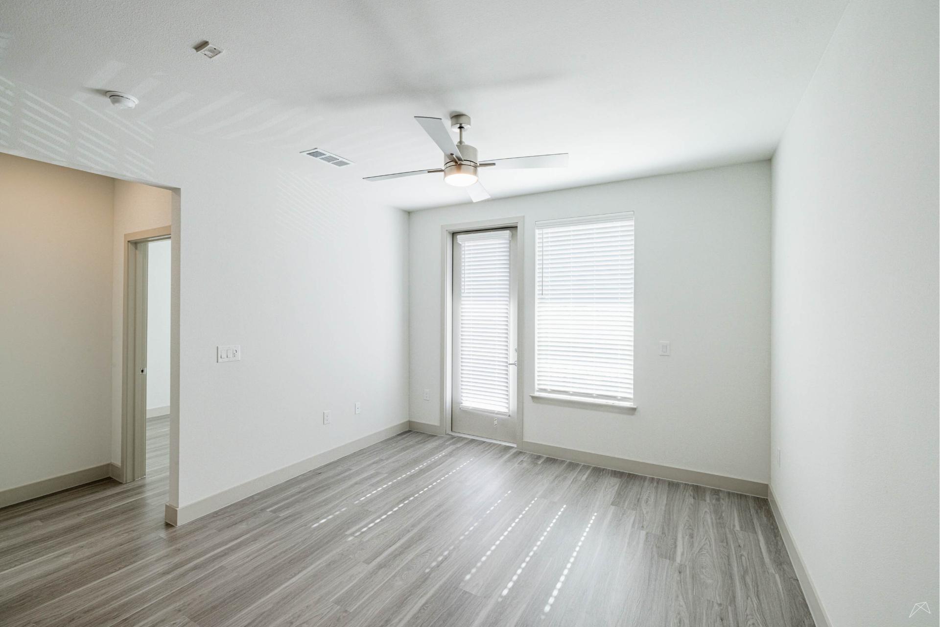 A bright, empty room with light wood-look flooring, white walls, a ceiling fan, two large windows with blinds, and a glass door outside.