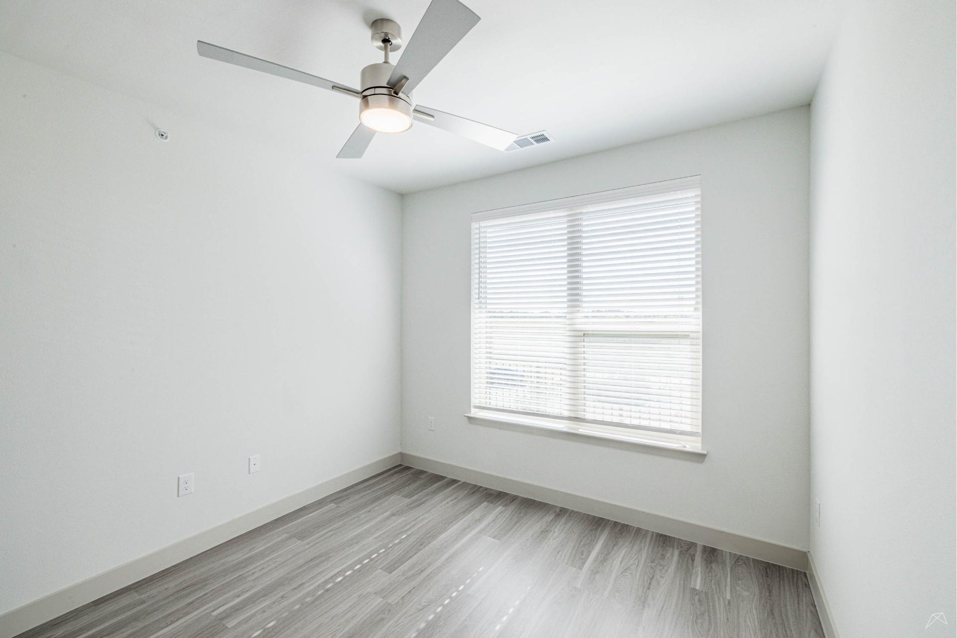 Bright, empty room with light gray wood-look flooring, white walls, large window with blinds, and a ceiling fan. Sunlight fills the space.