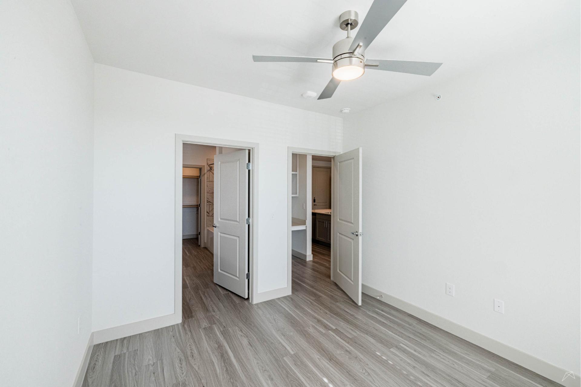 A bright, empty room with light-colored walls and wood-look flooring, a ceiling fan with light, and doors to a closet and hallway.