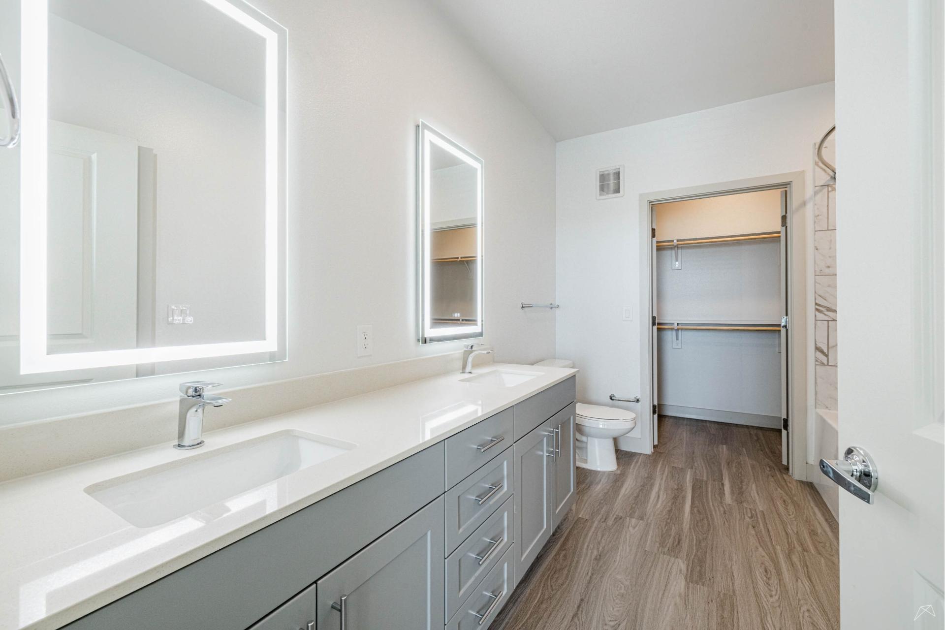 Modern bathroom with double-sink vanity, large lighted mirrors, wood-style flooring, open walk-in closet with shelves, and a toilet.