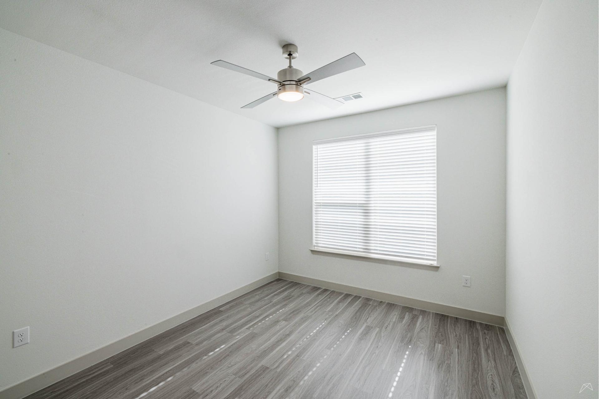 A bright, empty room with light gray walls, wood-look flooring, a large window with blinds, and a ceiling fan with a central light.