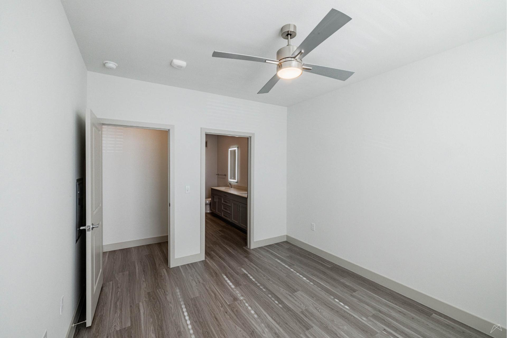 Empty, modern bedroom with gray wood-look flooring, white walls, ceiling fan with light, and open door to a bathroom with vanity and mirror.