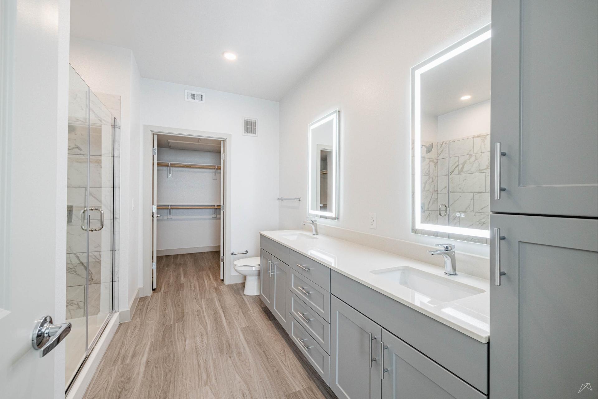 Modern bathroom with light gray cabinets, double sinks, large mirrors with built-in lighting, glass shower, toilet, wood-like flooring, and a walk-in closet in the background.
