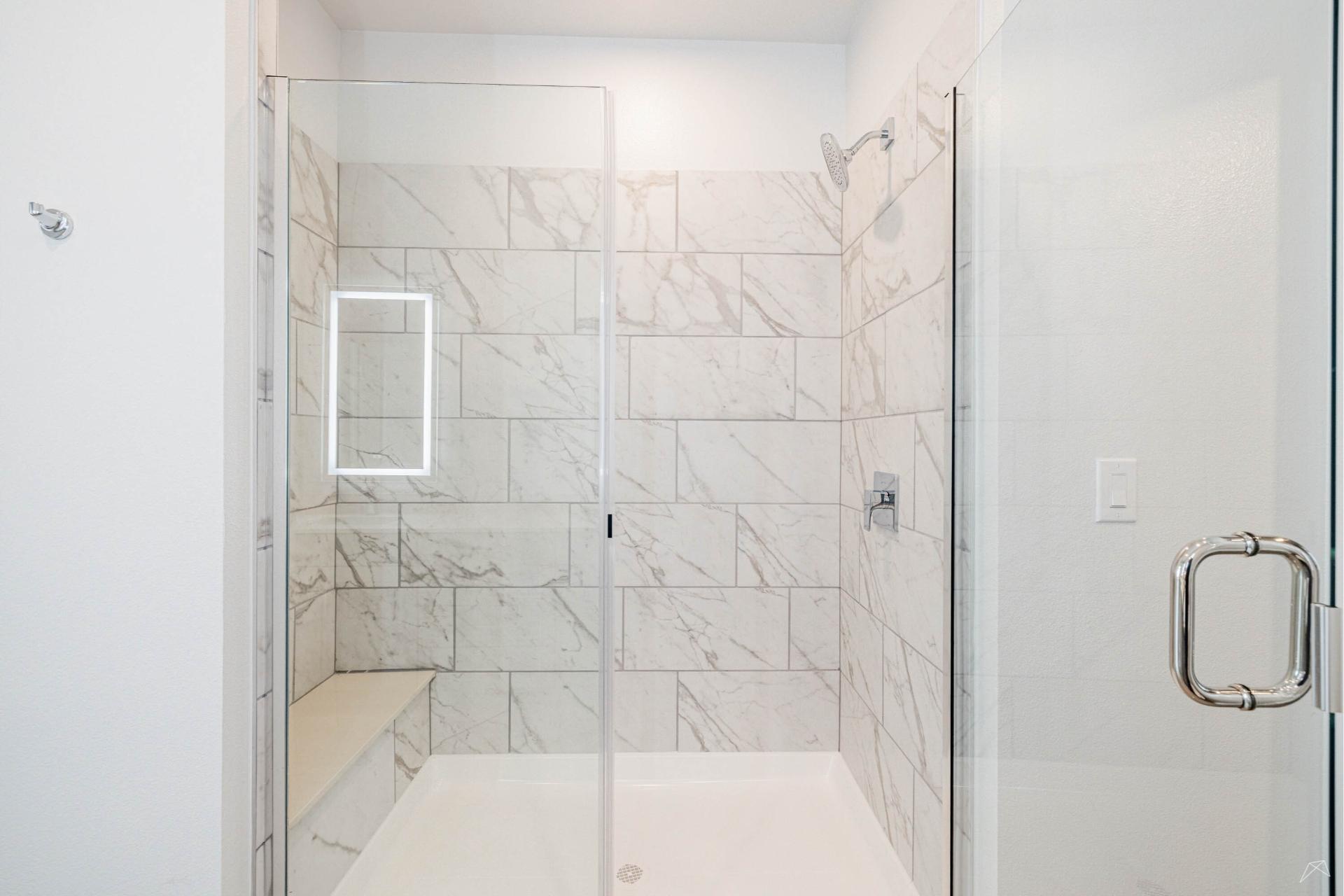 A modern walk-in shower with glass doors, marble tiles, a built-in bench, and wall-mounted showerhead. Lighted mirror outside.